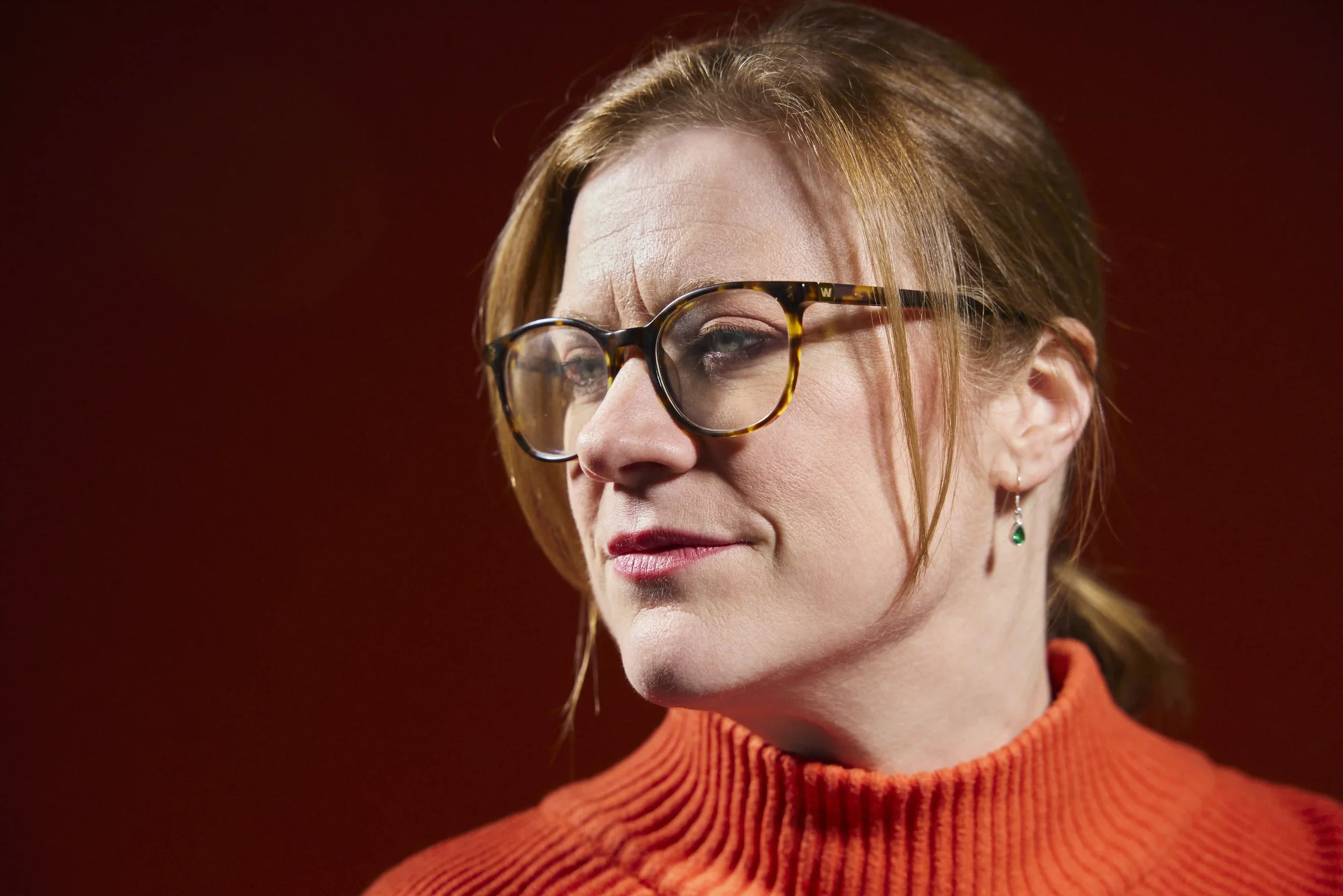 Close-up of a woman with red hair wearing tortoiseshell glasses and an orange turtleneck sweater, looking to the side against a dark red background.