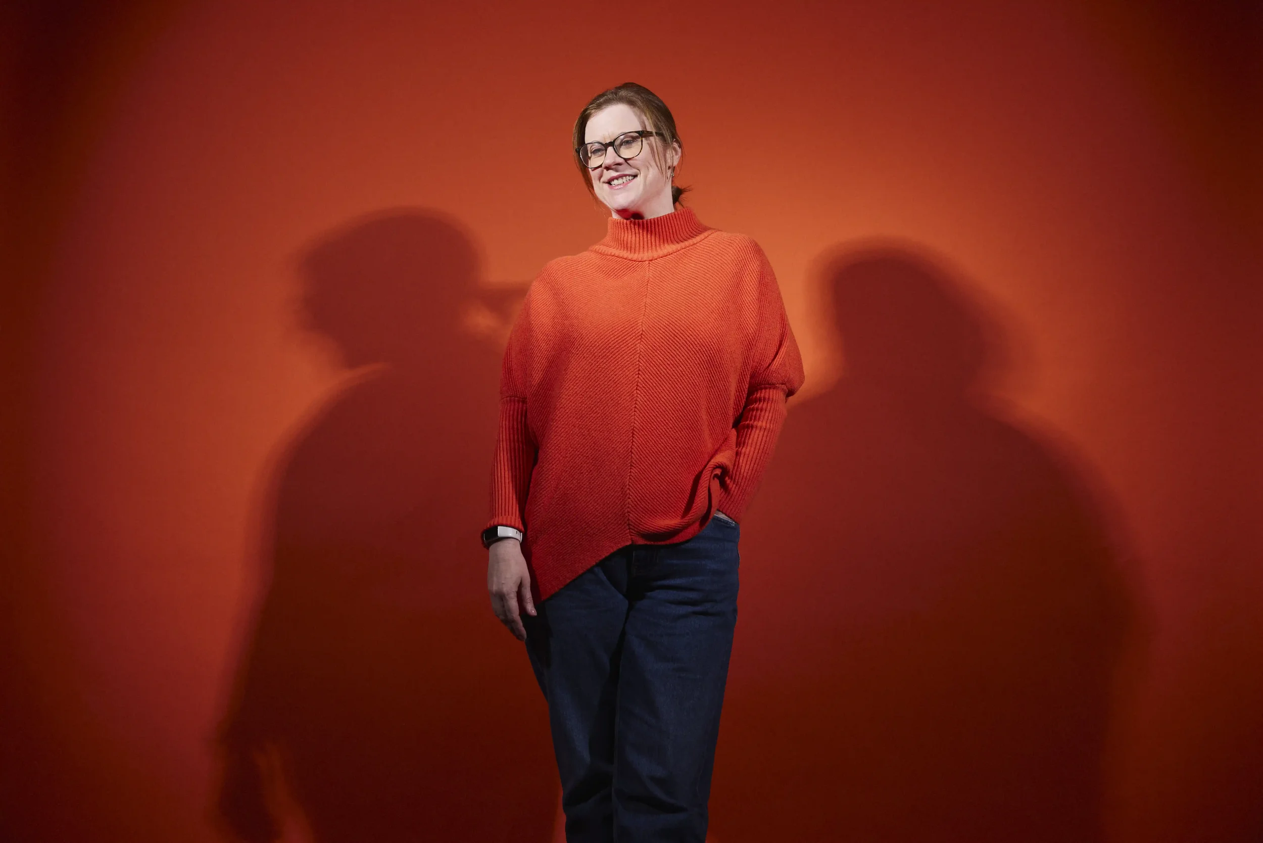 Woman with short hair, tied back, wearing glasses, a red turtleneck sweater, and dark pants, smiling while standing against a red background with blurred shadows of two other people.