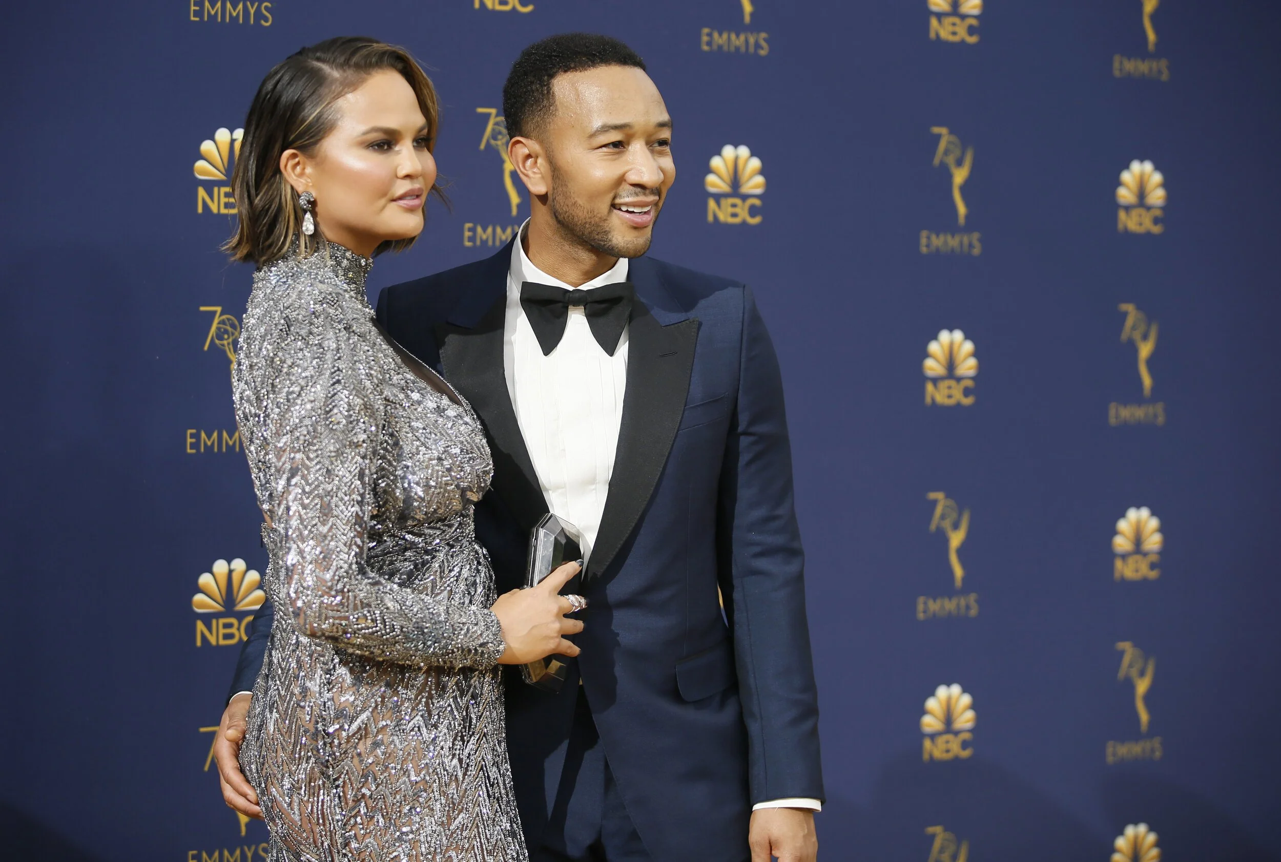 A woman in a silver, sequined gown and a man in a navy tuxedo with a black bow tie pose together at an Emmy Awards event with a dark blue backdrop featuring NBC and Emmy logos.
