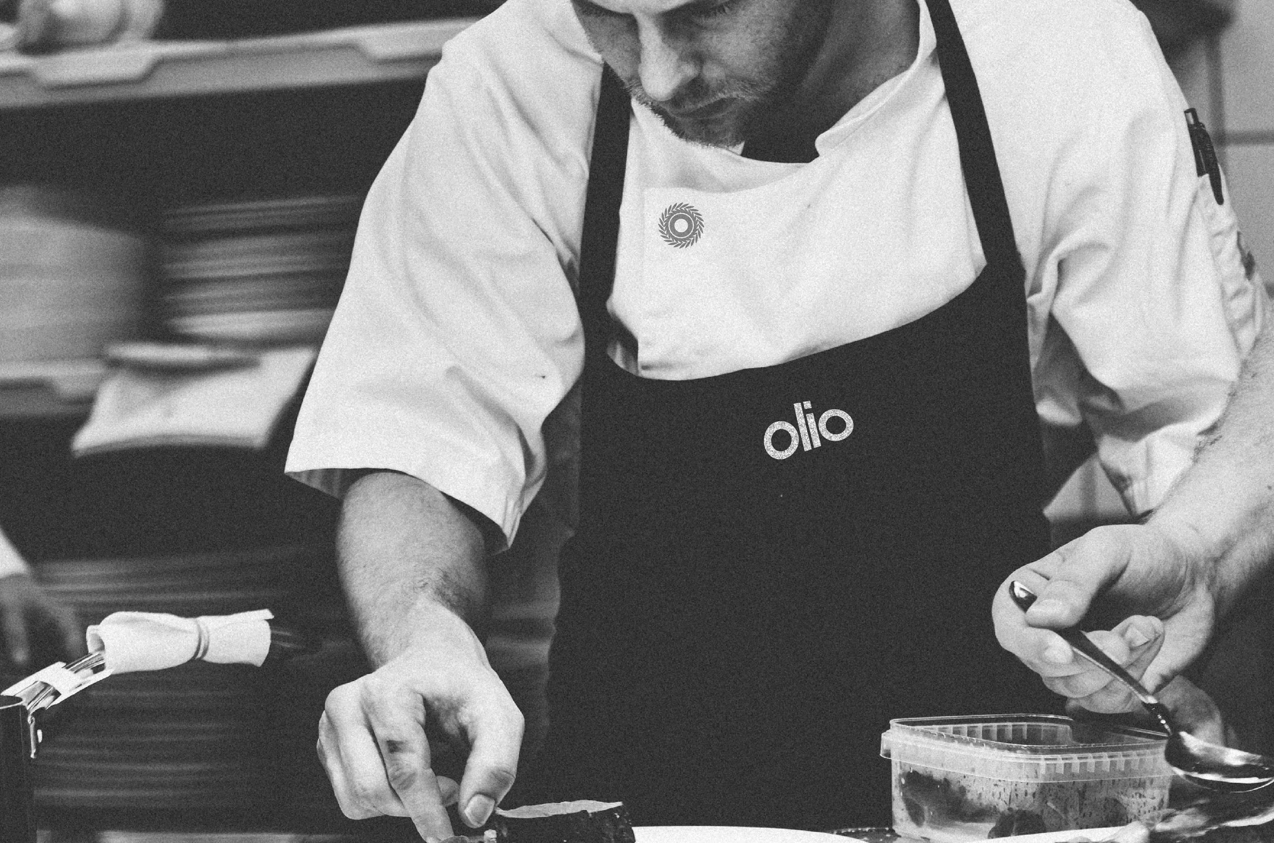 A chef with a beard wearing a white chef's coat and a black apron with the word 'olio' on it, preparing food in a kitchen. The chef is holding a spoon and working over a plate.