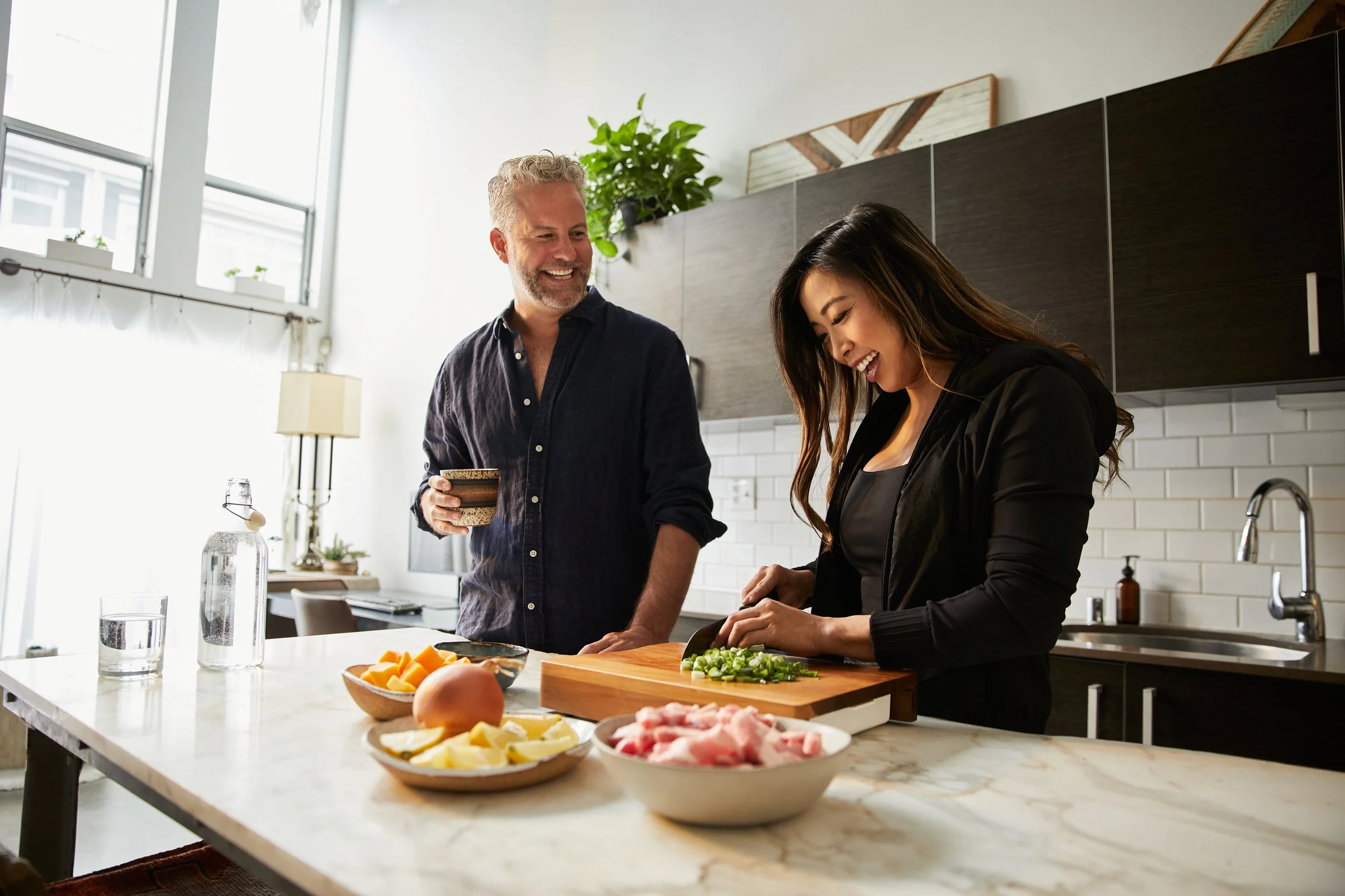 A man and woman cooking together in a modern kitchen, the woman chopping green onions at a white marble counter, with bowls of sliced fruit and raw meat around them, and a large window letting in natural light.