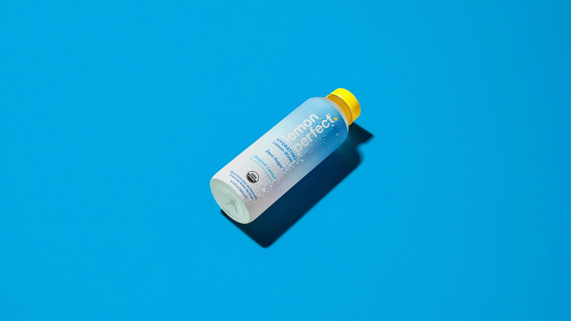 A bottle of Lemon Perfect hydration water with a yellow cap on a blue background, with a shadow cast to the right.