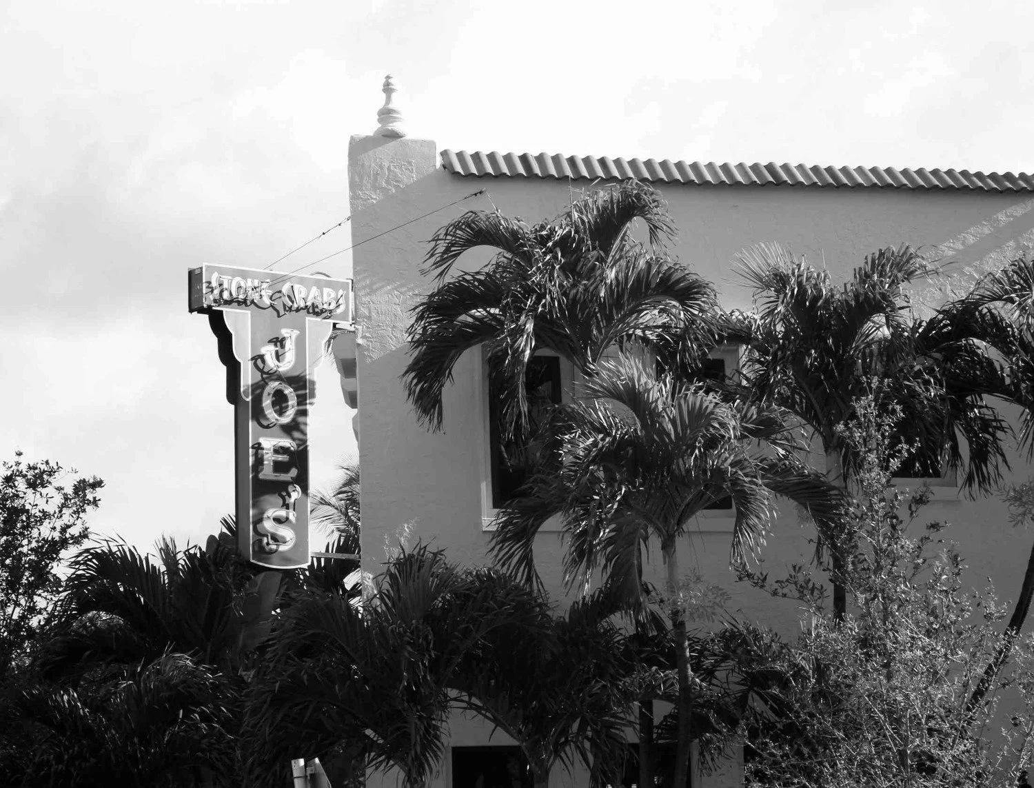 A black and white photo of a building with a restaurant sign that reads 'Home of the Crab'. The building has a tiled roof and is partially obscured by palm trees.