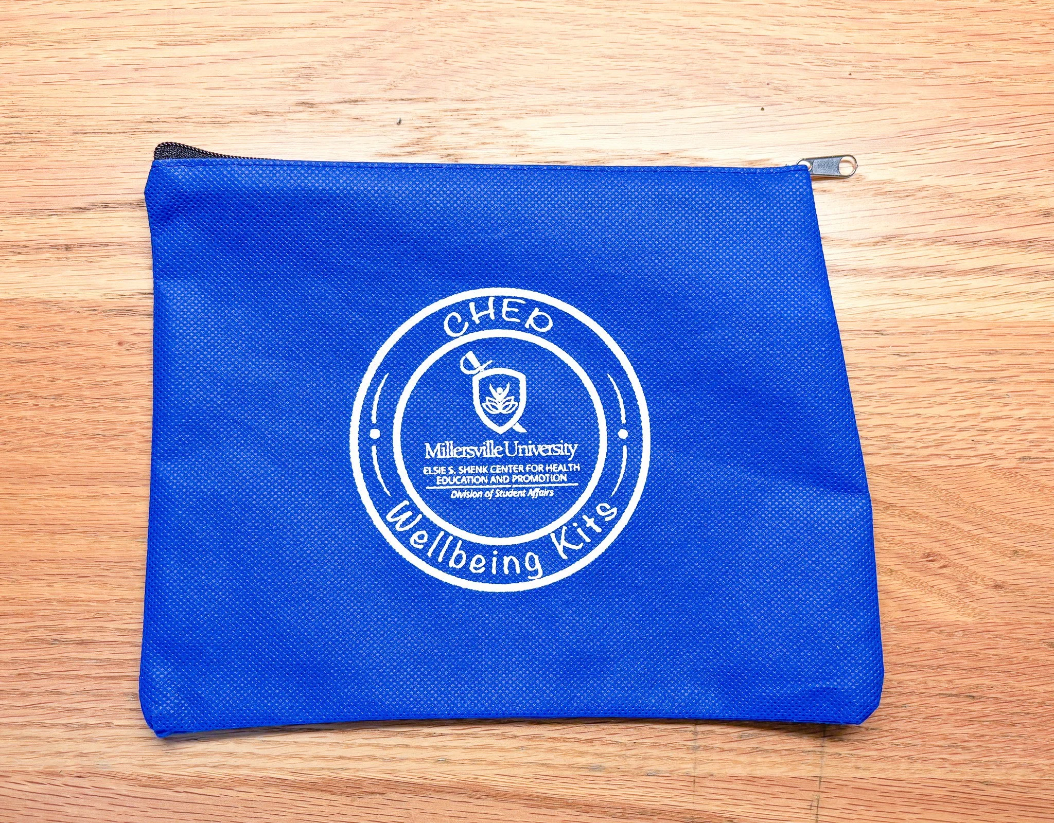 Blue Zippered pouch with white logos and text, placed on a wooden surface. The logo includes a leaf and the words 'CHEQ,' 'Millersville University,' and 'Wellbeing Kits'.
