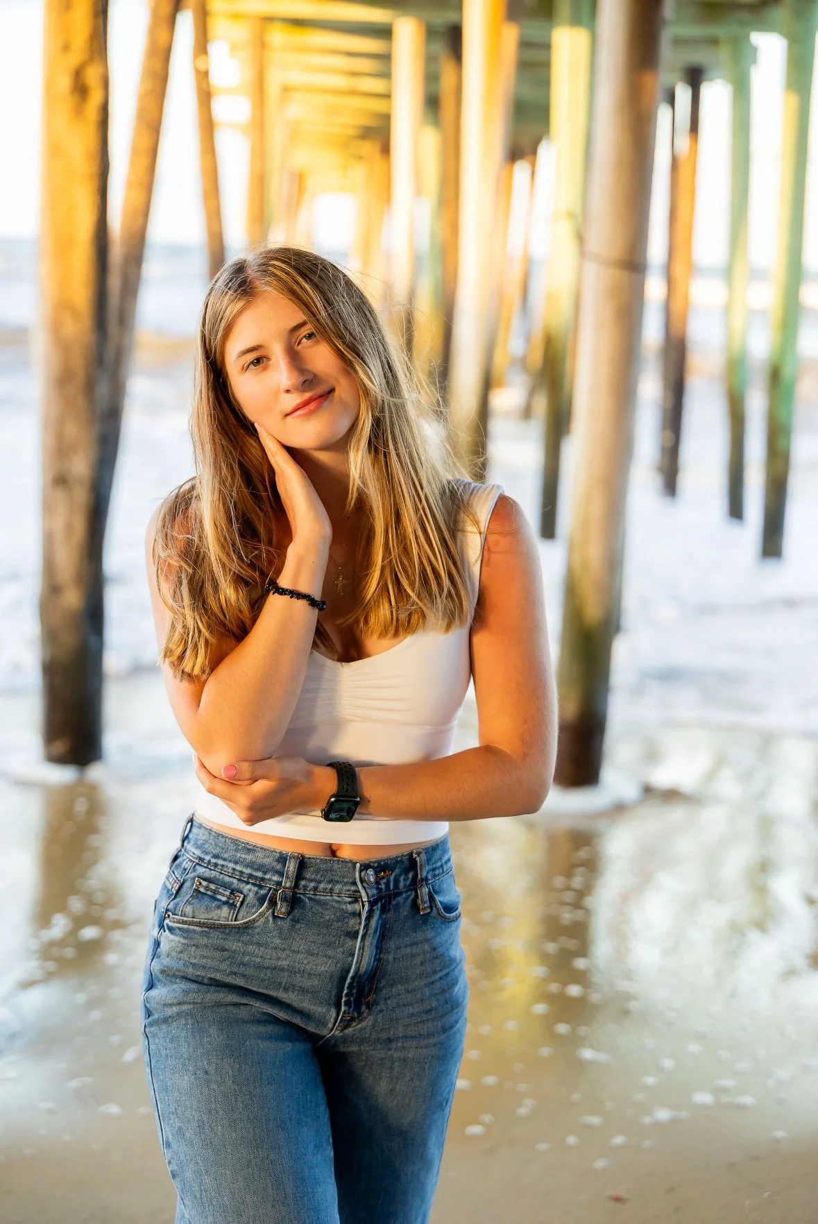 A young woman standing under a pier at the beach, with her hand touching her face, wearing a white tank top, jeans, and a black watch, smiling softly at the camera.