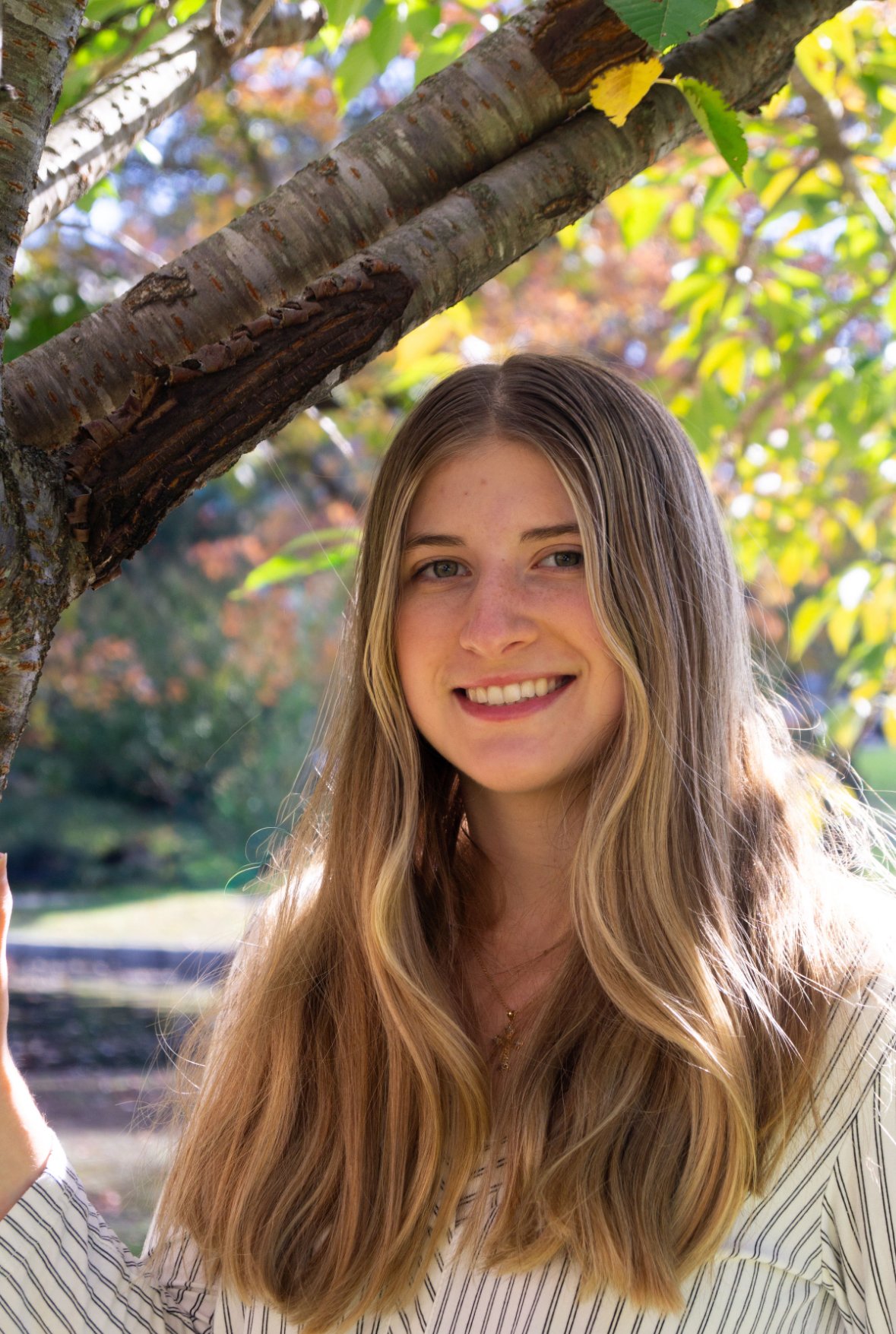 A young woman with long blonde hair smiling outdoors near a tree with green leaves and colorful autumn foliage in the background.