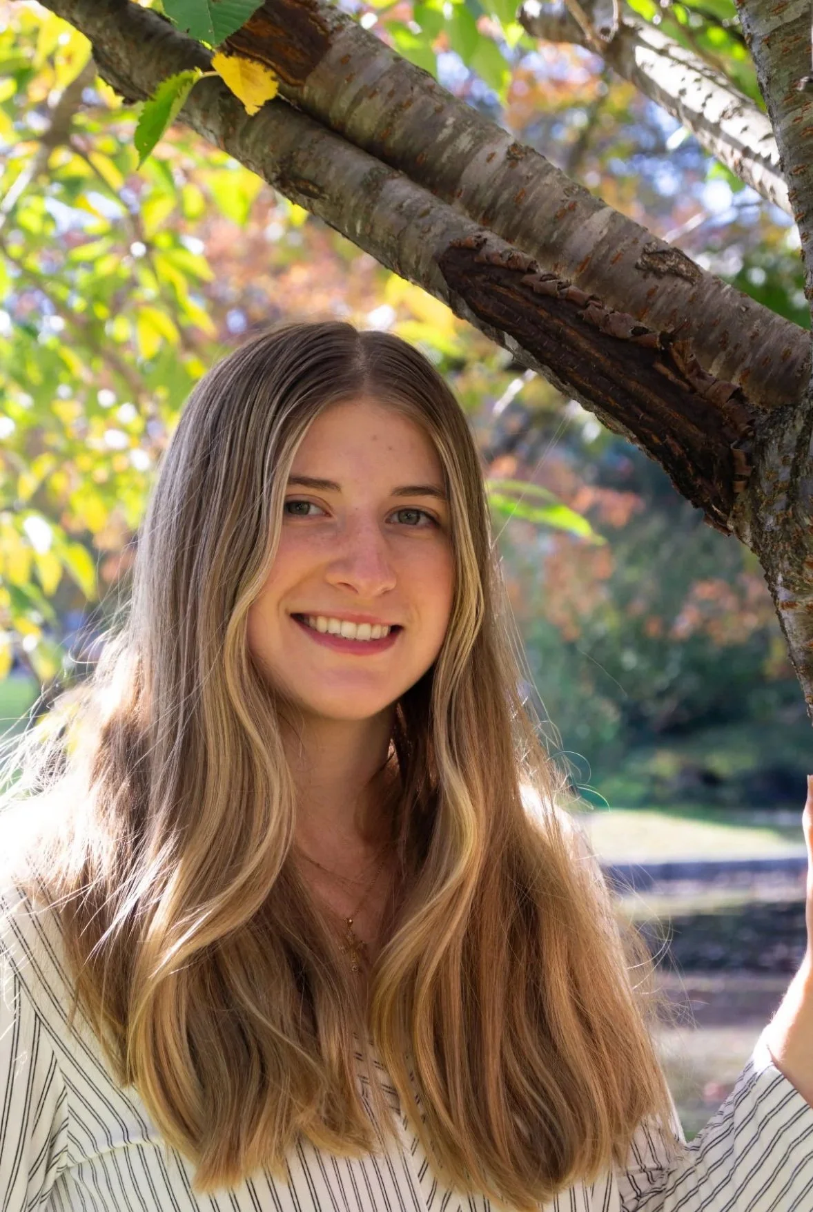 A young woman with long, wavy blond hair smiling outdoors near a tree with green and orange leaves.