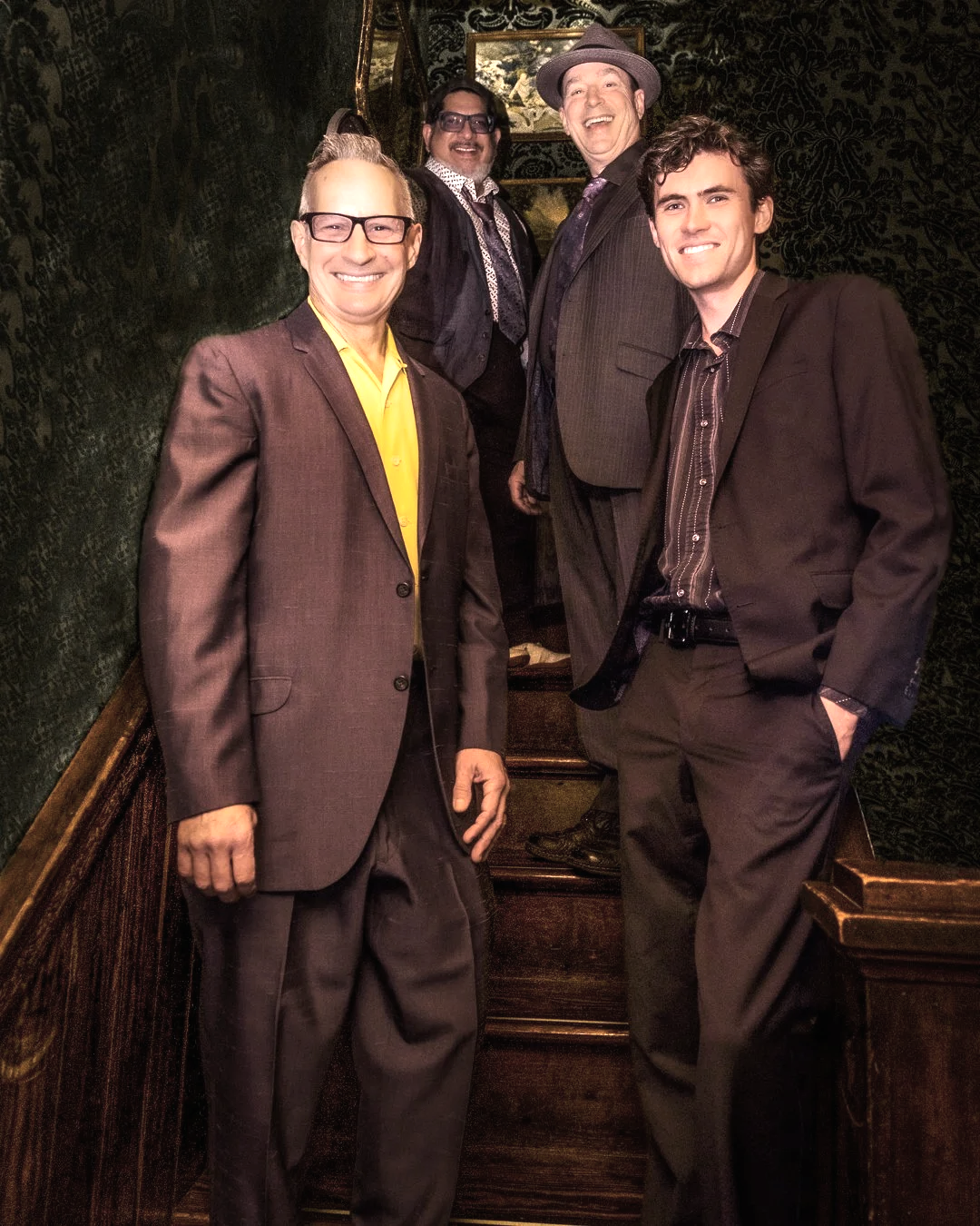 A group of five men in suits posing on a staircase inside a dimly lit, elegant room with dark ornate wallpaper. They are smiling and appear to be in a cheerful mood.