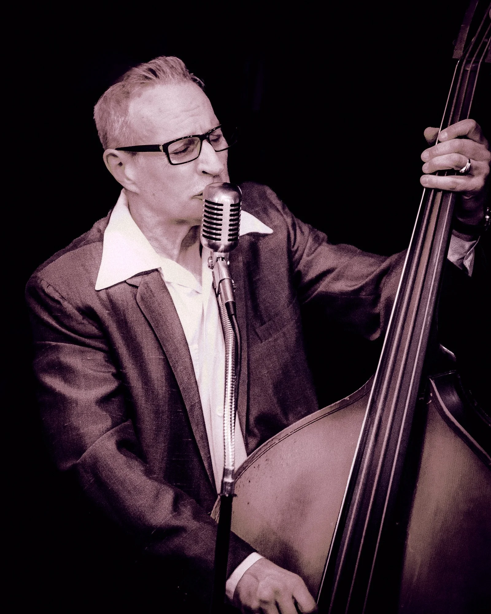 A man with glasses wearing a suit and white shirt playing double bass and singing into a vintage microphone against a dark background.