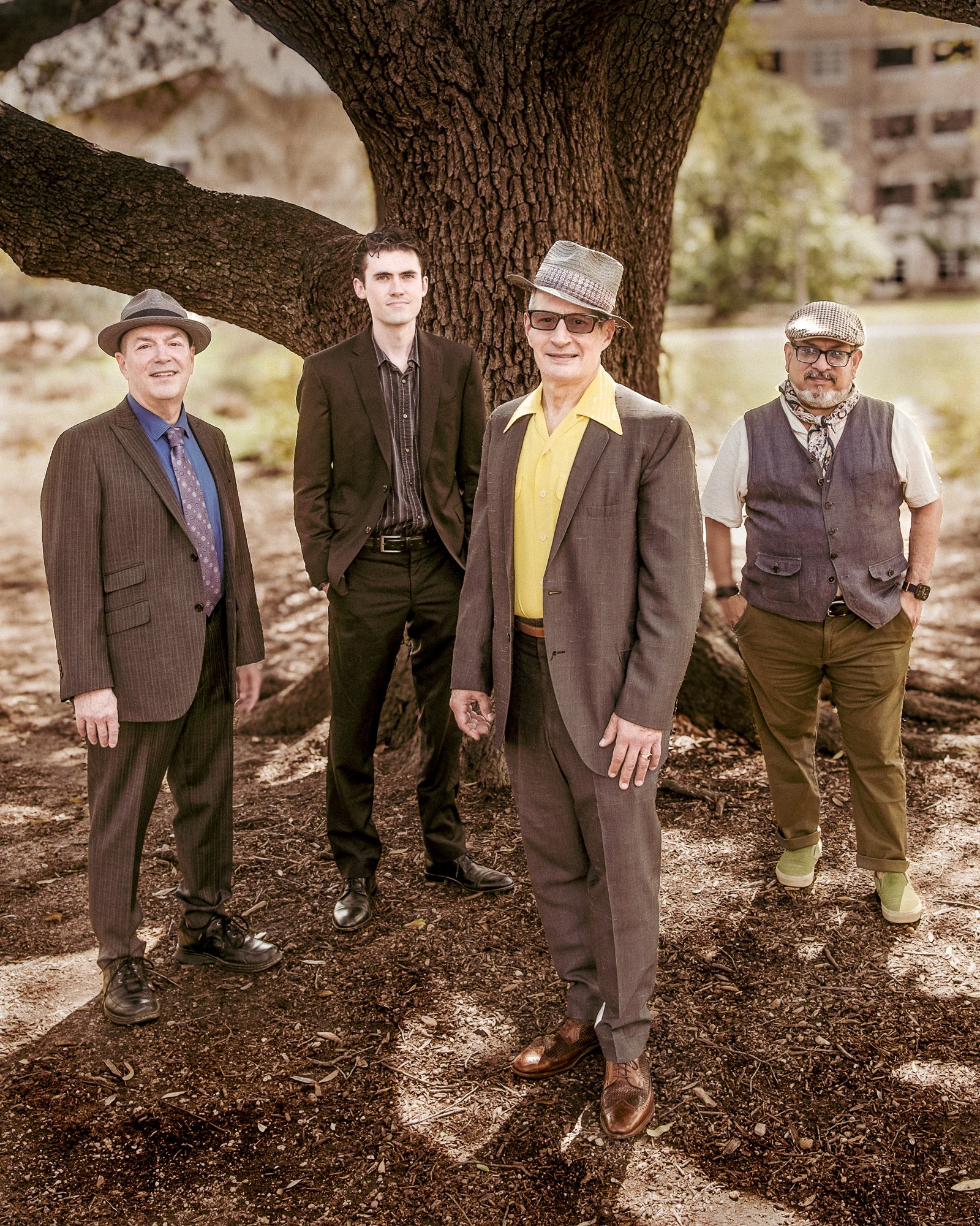 Four men standing outdoors in front of a large tree. They are dressed in various styles of suits and casual wear, with some wearing hats and glasses.