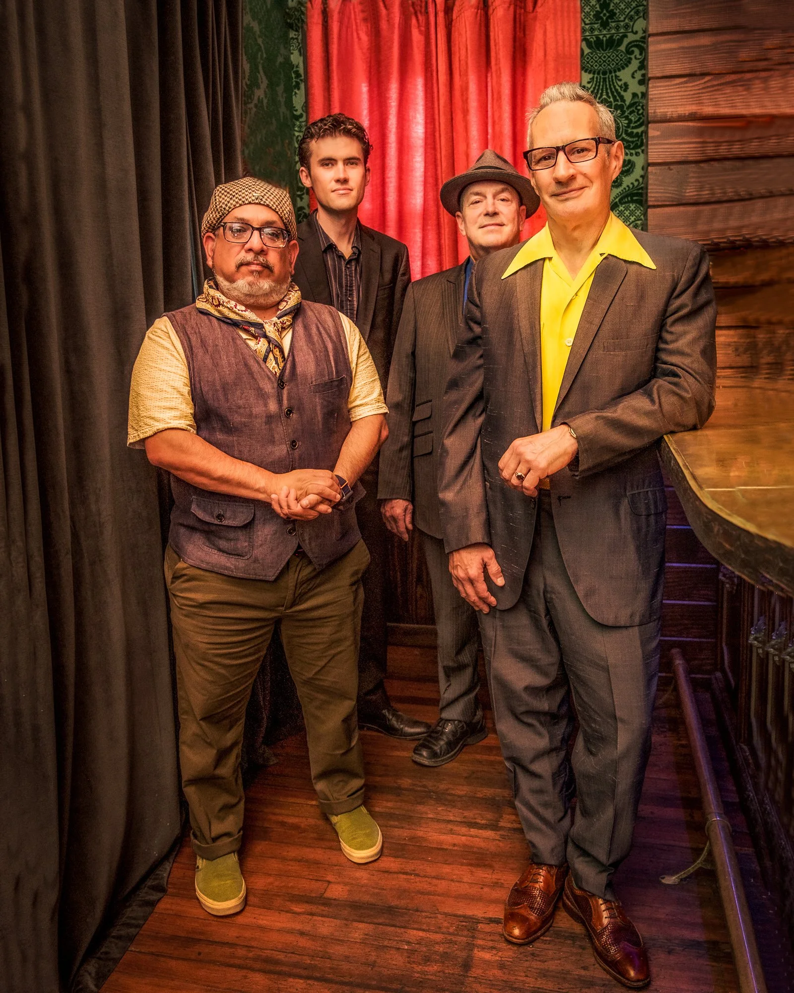Four men standing together in a room with wooden floors, red curtains, and dark-colored walls with a green patterned wall detail.