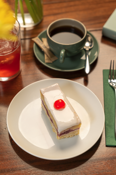 Bakewell Slice, a twist on a classic with a buttery shortbread base, layer of raspberry jam, almond frangipane filling and a nostalgic thick icing topped with a glace cherry.