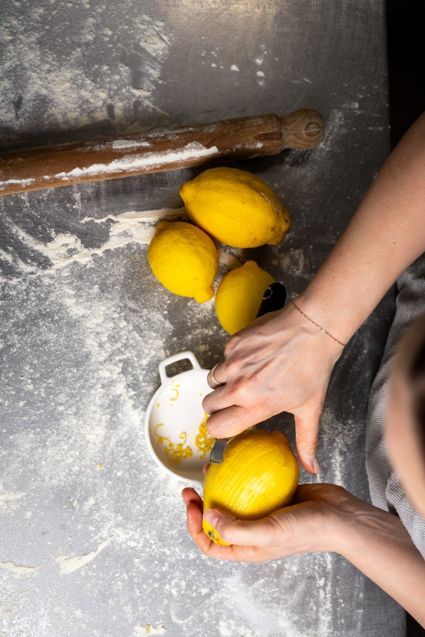 Person peeling a lemon in a kitchen with a flour-dusted surface, a rolling pin, and several whole lemons.