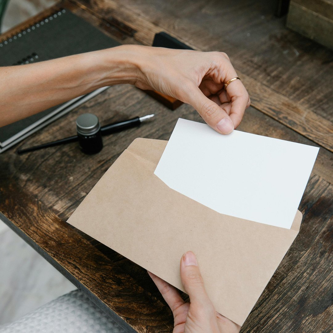 Eine Person hält einen Briefumschlag mit einem weißen Brief darin, sitzend an einem Holztisch.