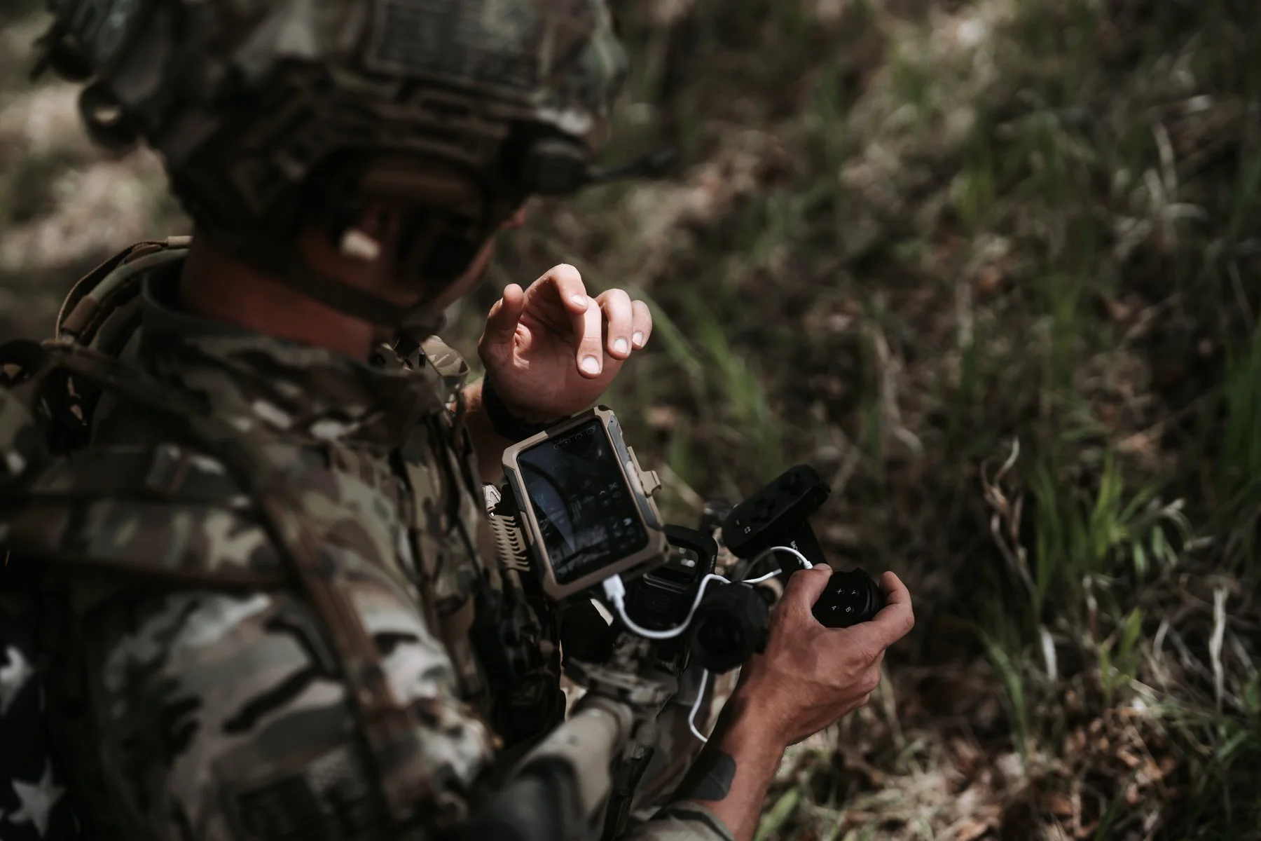 Soldier in camouflage uniform operating a drone controller in a grassy outdoor environment.