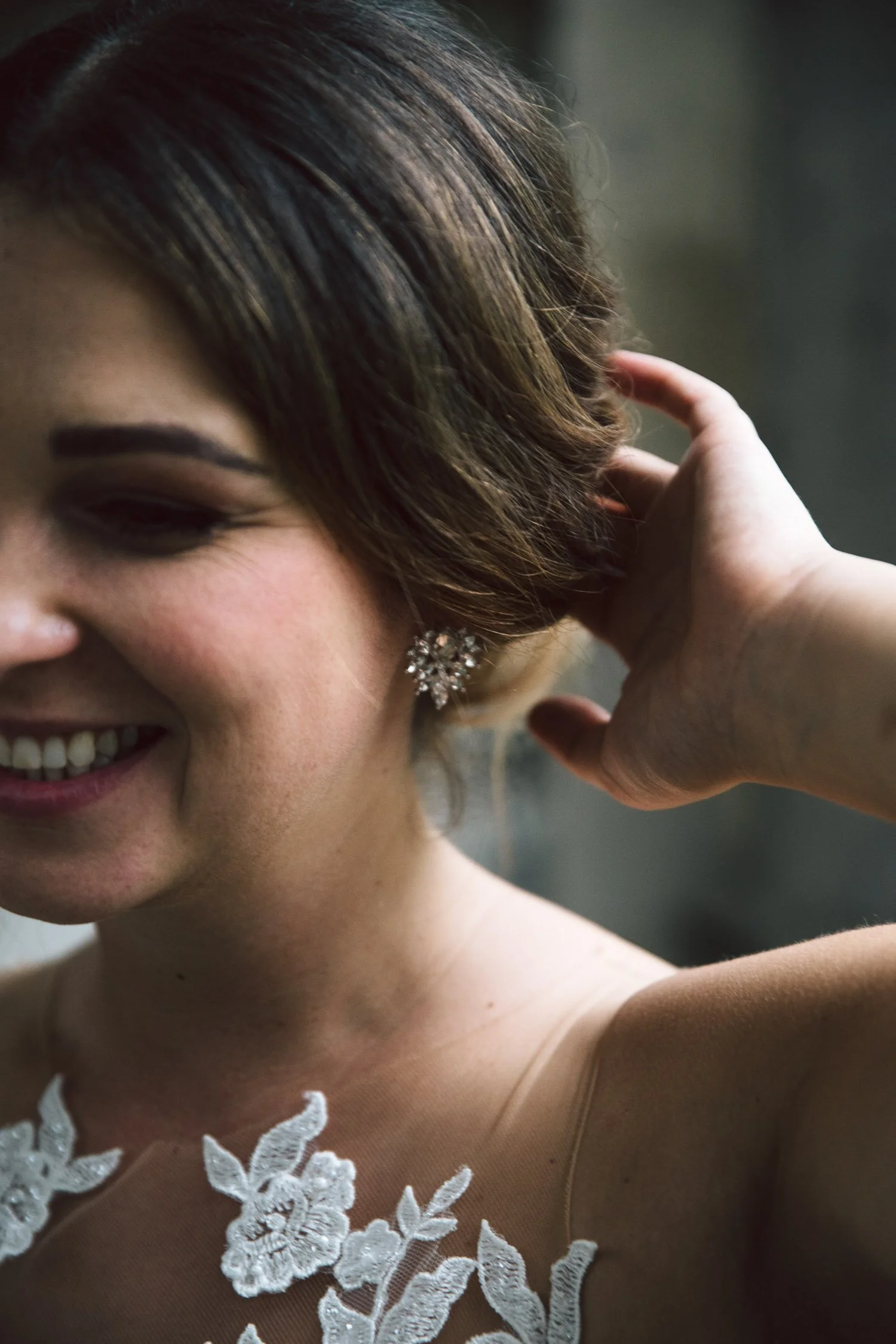 Nahaufnahme einer lächelnden Frau mit braunen, gewellten Haaren, die ein elegantes Kleid mit Blumenstickerei trägt, und einem funkelnden Ohrring, während sie ihre Hand an den Hinterkopf legt.