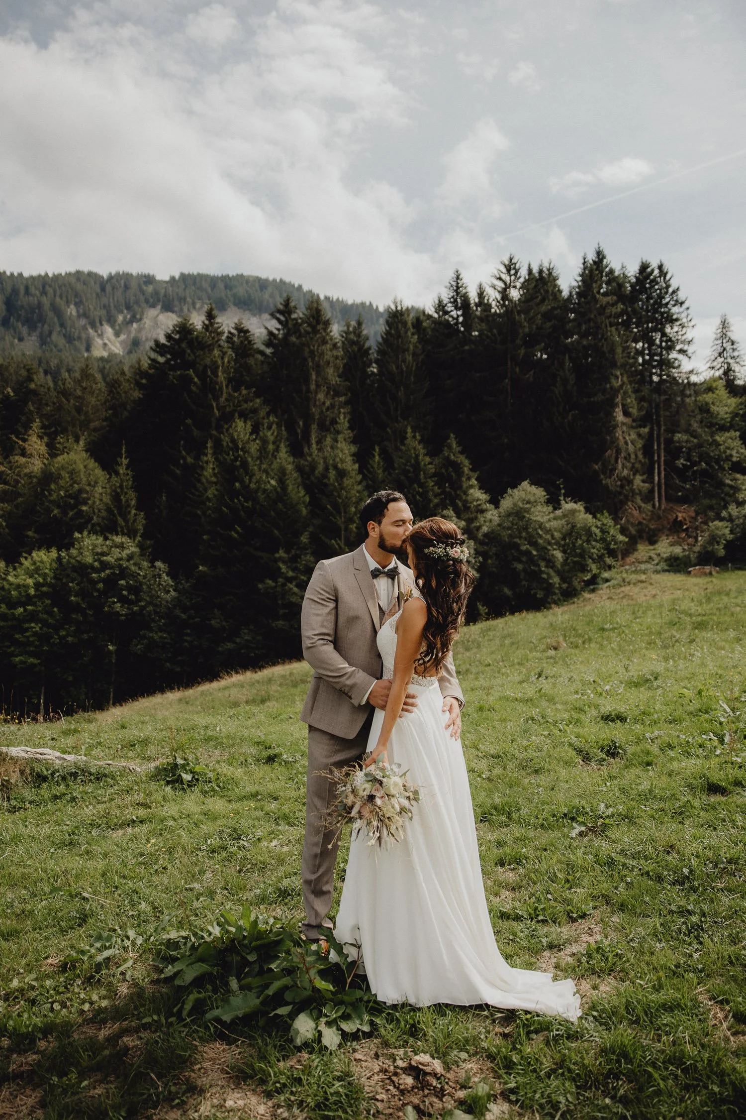 Braut und Bräutigam küssen sich auf einer Wiese mit einem Wald im Hintergrund, bei einer Hochzeit im Freien.