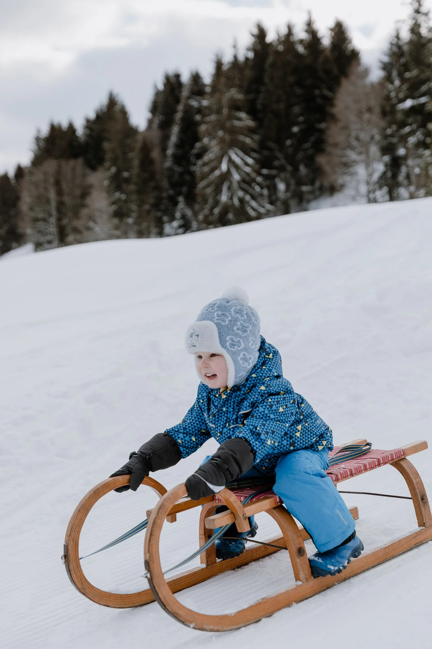 Ein lachender kleiner Junge in Winterkleidung fährt auf einem Holzschlitten durch den Schnee.