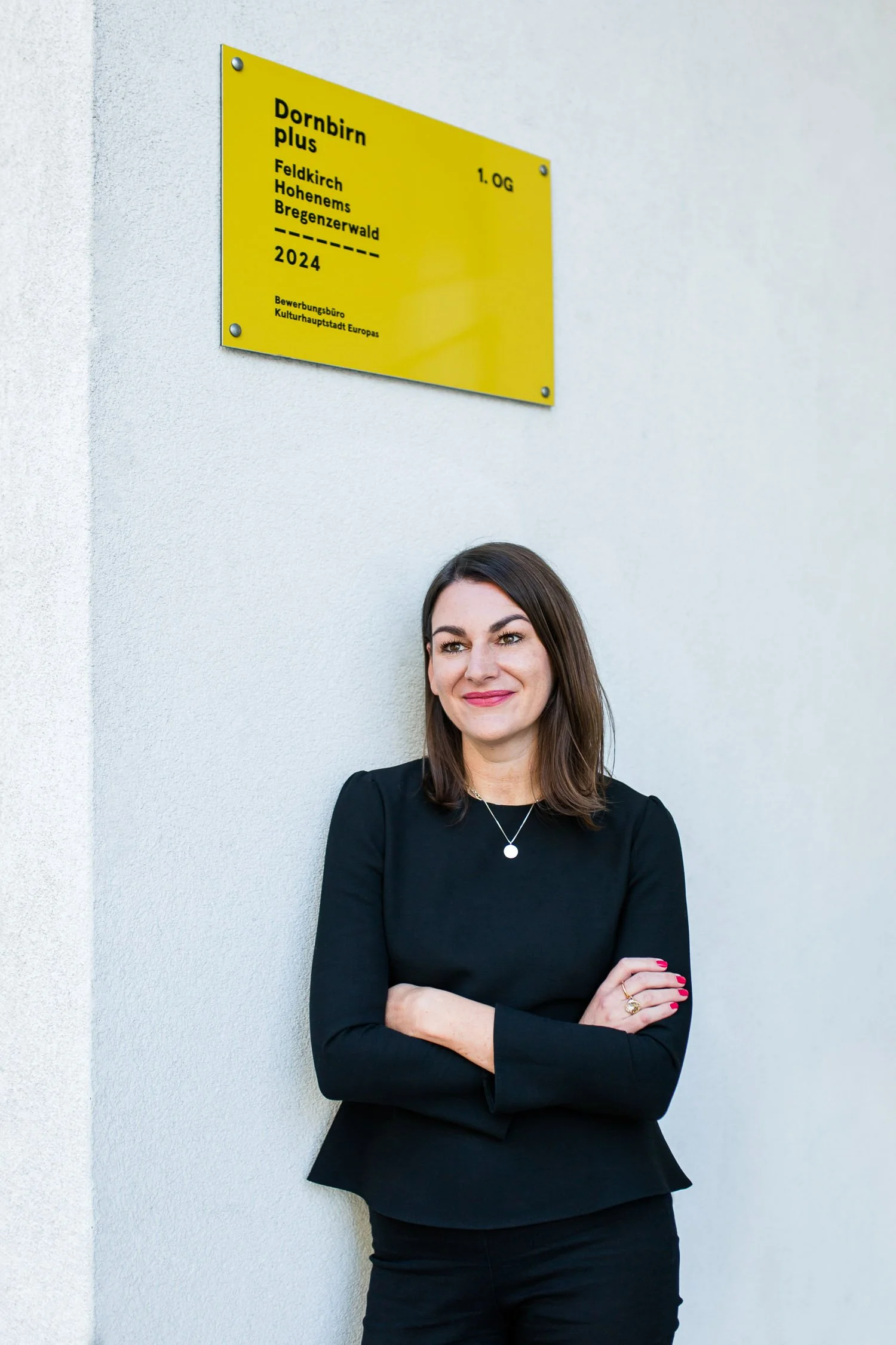 Eine Frau steht vor einer weißen Wand mit einem gelben Schild, auf dem Standorte und das Jahr 2024 abgebildet sind, und lächelt.