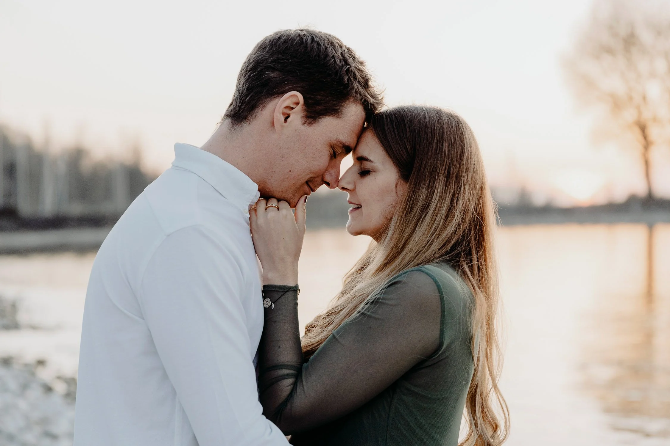 Ein verliebtes Paar steht eng beieinander am Wasser bei Sonnenuntergang, mit gesenkten Stirn und geschlossenen Augen, in einer intimen Geste.