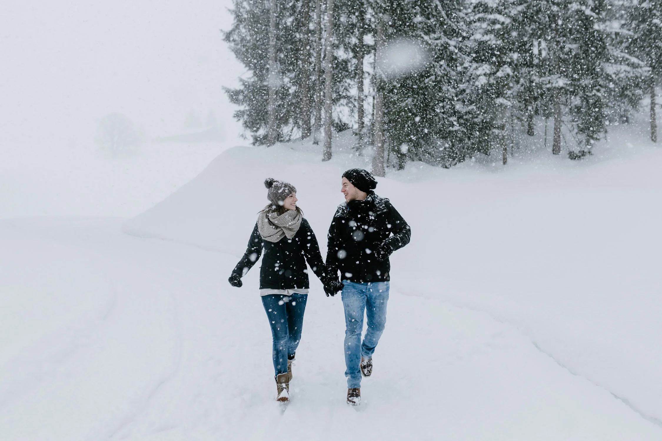 Ein Paar läuft im Schneesturm hand in hand durch eine verschneite Landschaft mit Bäumen im Hintergrund.