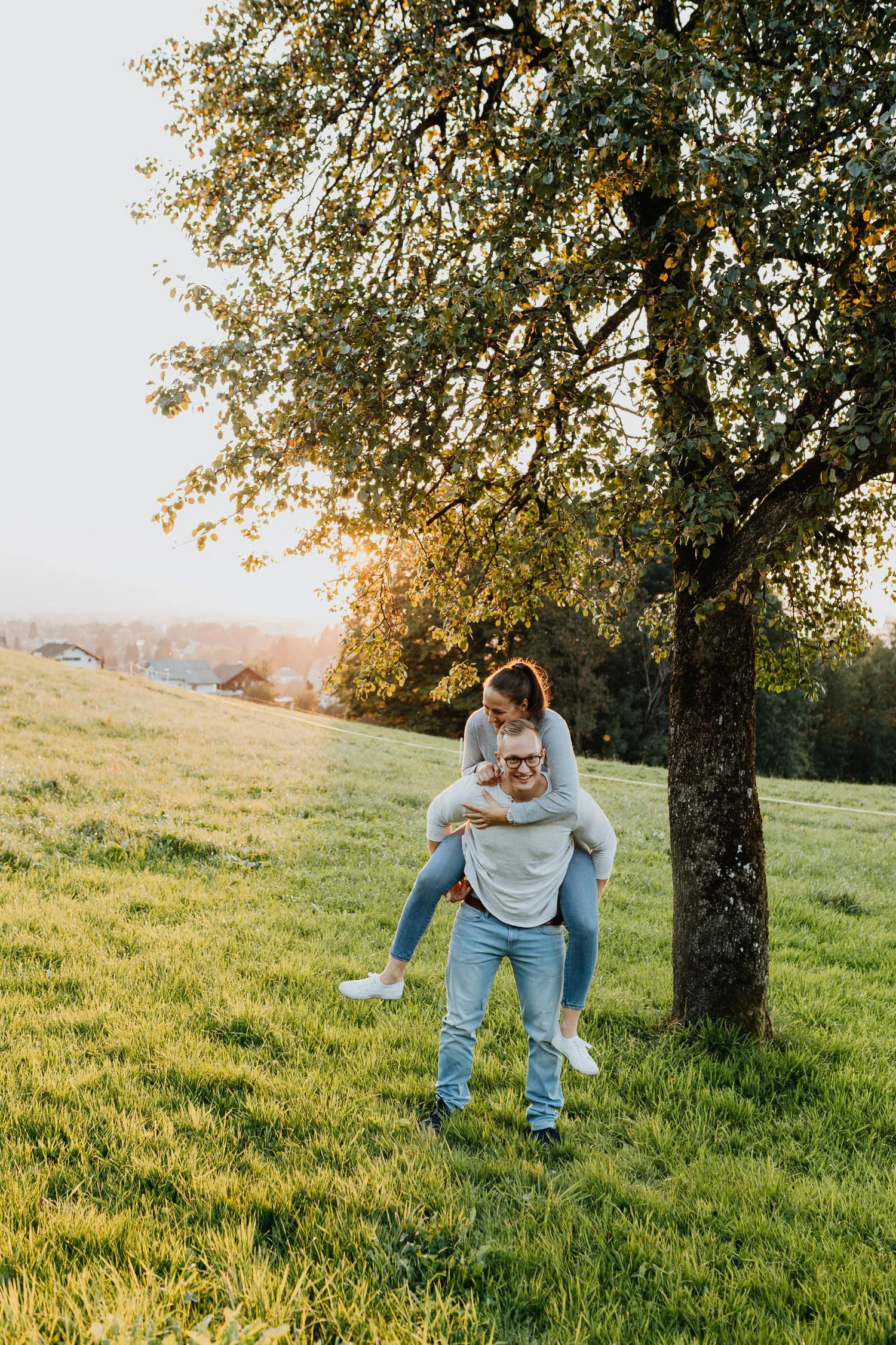 Ein Paar, das im Freien unter einem Baum steht, während die Frau auf dem Rücken des Mannes sitzt, in einer ländlichen Gegend bei Sonnenuntergang.