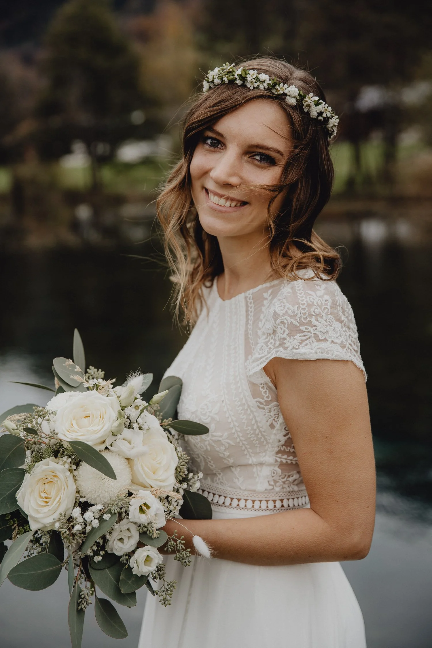 Eine Frau trägt ein weißes Hochzeitskleid und einen Blumenkranz im Haar, hält einen Brautstrauß aus weißen Rosen und Grünblättern, steht vor einem Wasser im Freien.