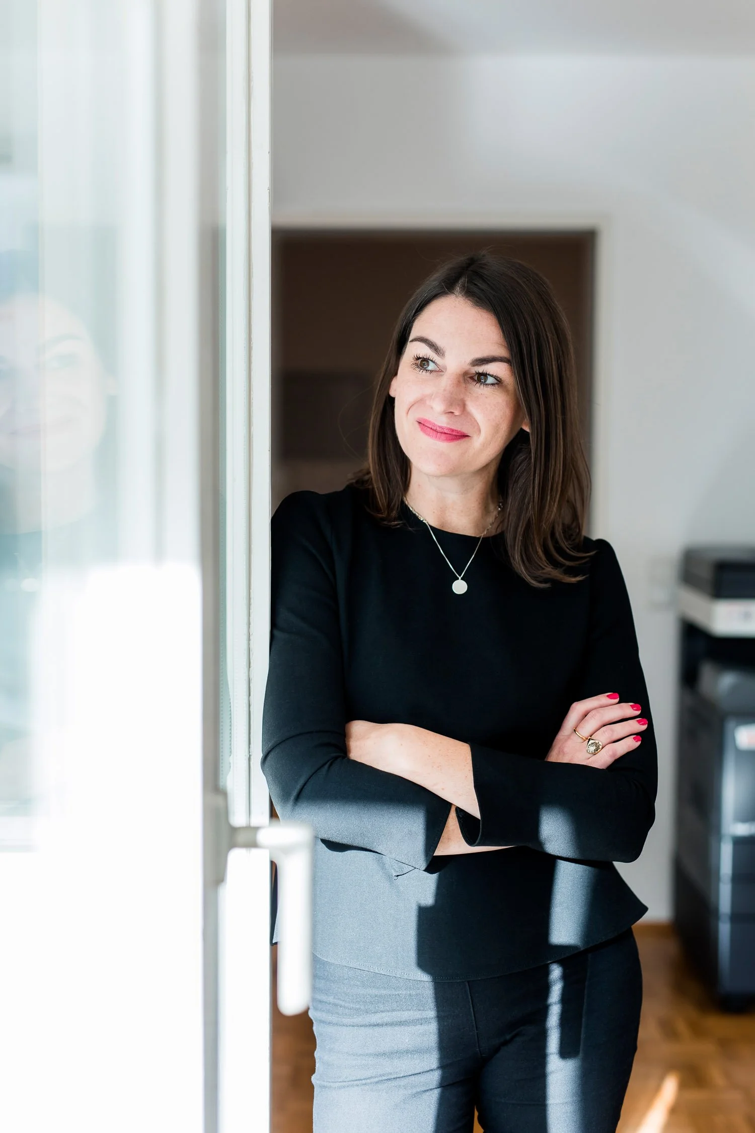 Eine Frau mit schwarzen Haaren und rotem Lippenstift, die im Büro neben einer Tür steht, mit verschränkten Armen und einem Lächeln, im Hintergrund ein Drucker.
