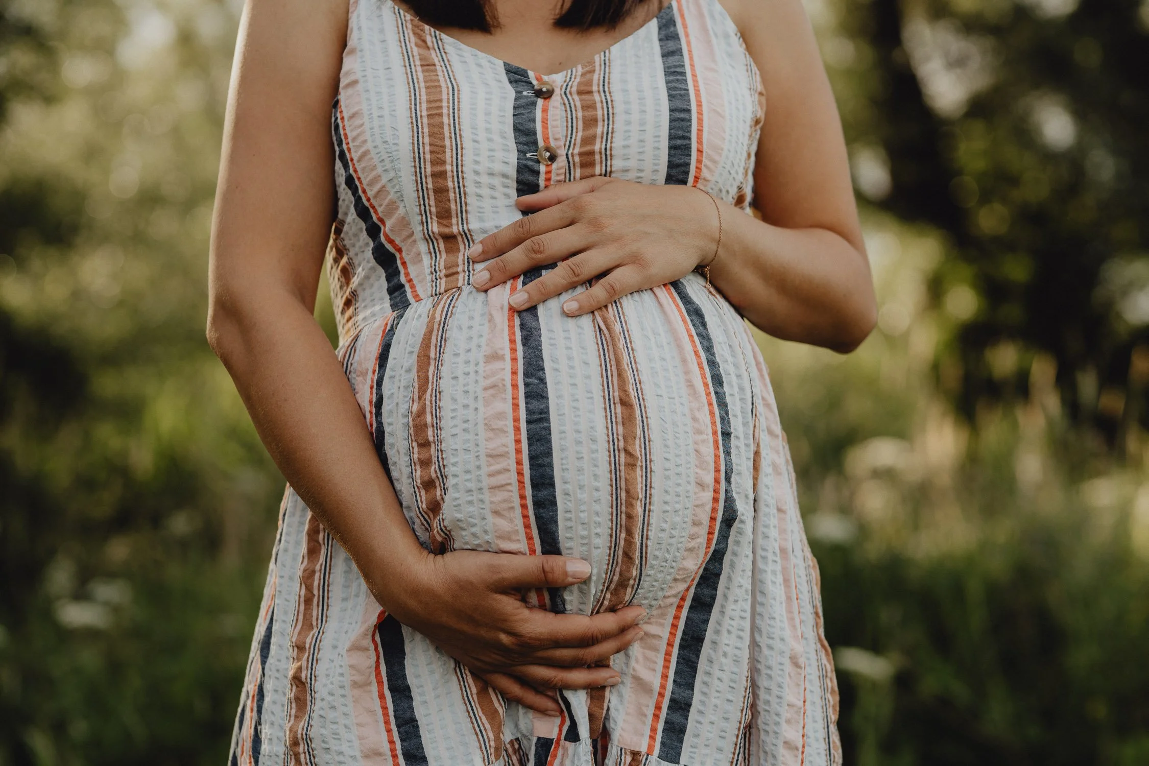 Nahaufnahme eines schwangeren Frauenbauchs, gehalten von ihren Händen, draußen mit grünem Hintergrund.