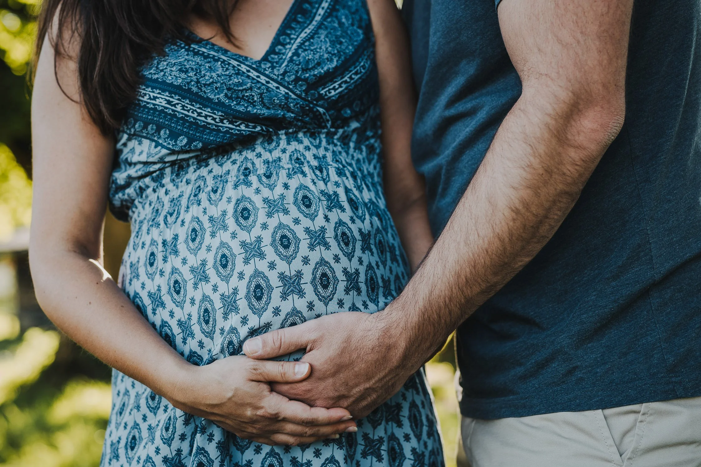 Ein schwangere Frau in einem blauen kleid hält die Hände ihres Partners auf ihrem Bauch. Die Szene spielt im Freien.