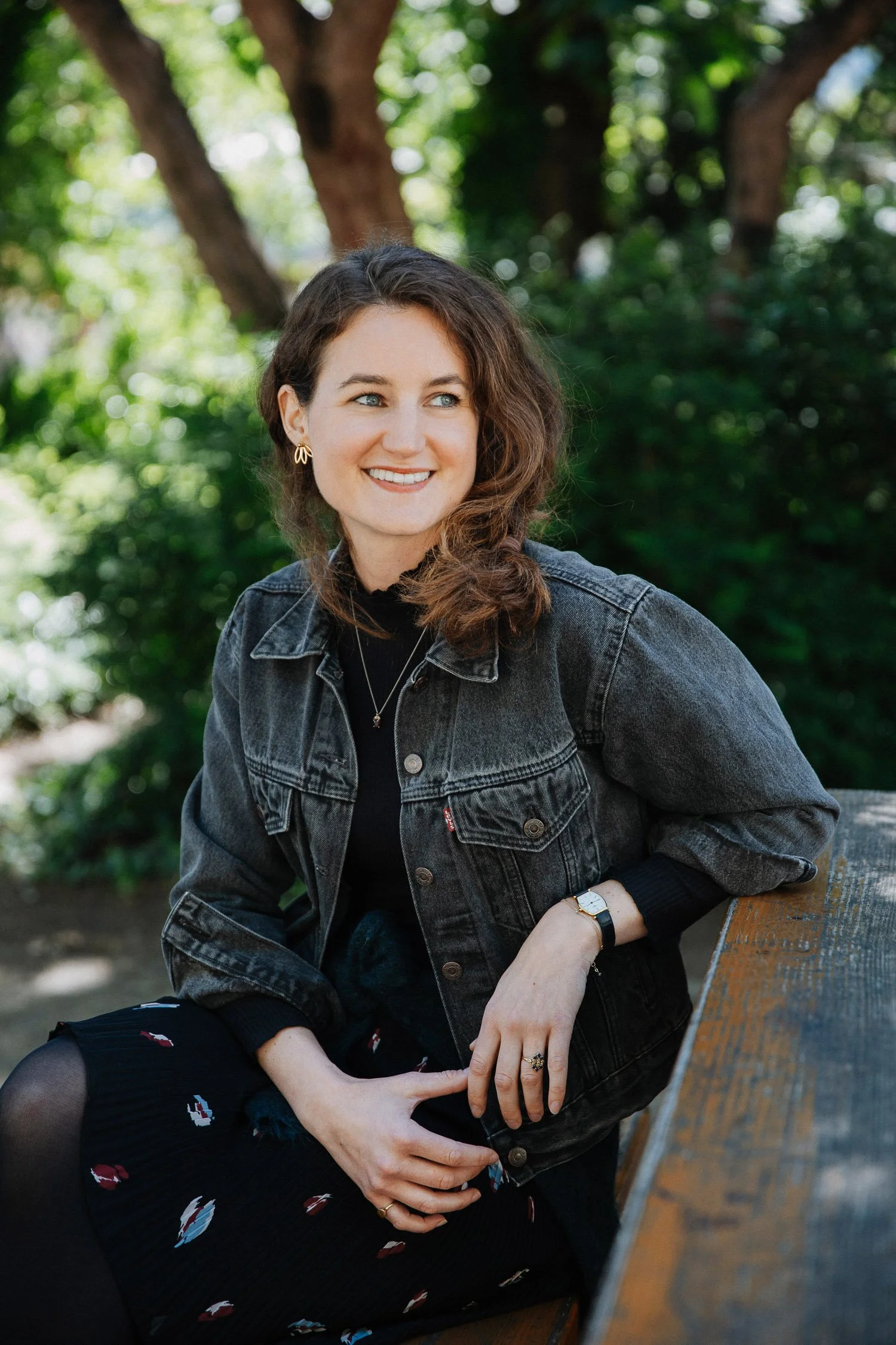 Jugendliche Frau mit braunen, gewellten Haaren sitzt auf einer Parkbank, trägt eine schwarze Jacke und ein schwarzes Kleid mit bunten Mustern, im Hintergrund grüne Bäume.