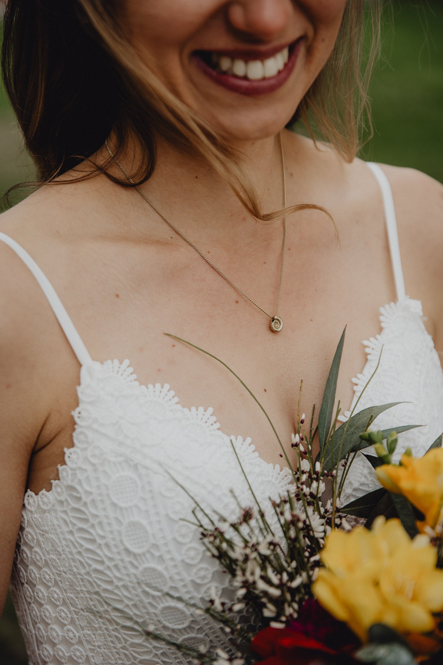 Eine Frau trägt ein weißes Spitzenkleid und hält einen Blumenstrauß, trägt eine Halskette, und lächelt. 