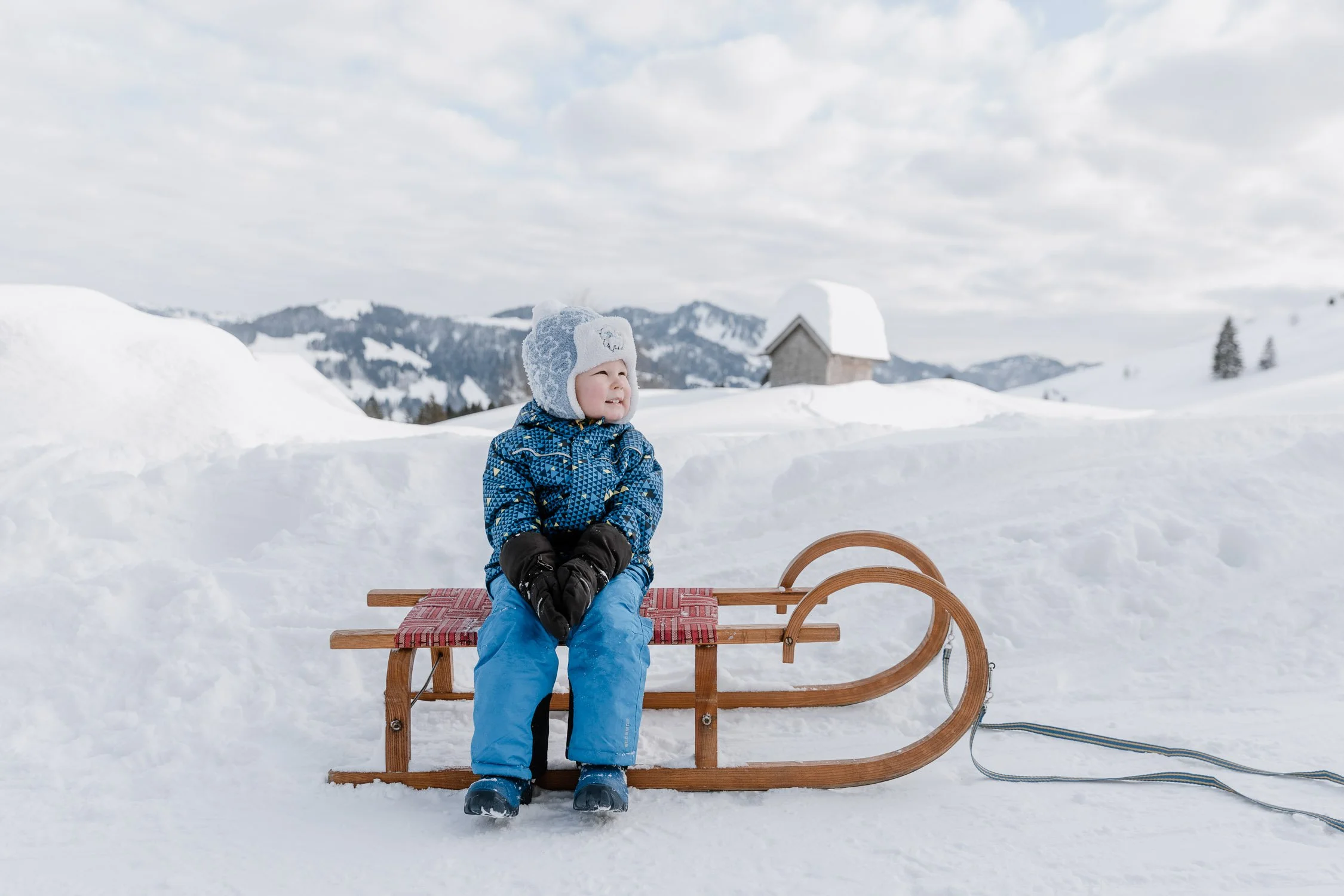 Ein Kind sitzt auf einem Schlitten im Schnee in einer winterlichen Landschaft mit Bergen und einem Haus im Hintergrund.