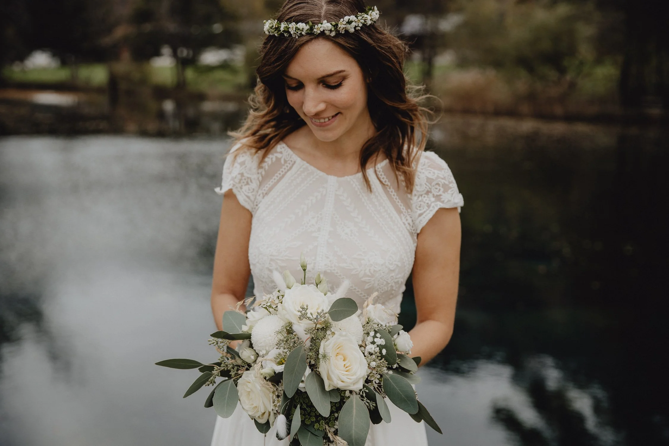 Braut in weißem Kleid mit Blumenkranz im Haar, die einen weißen Blumenstrauß hält, steht an einem Wasser, im Hintergrund Bäume.