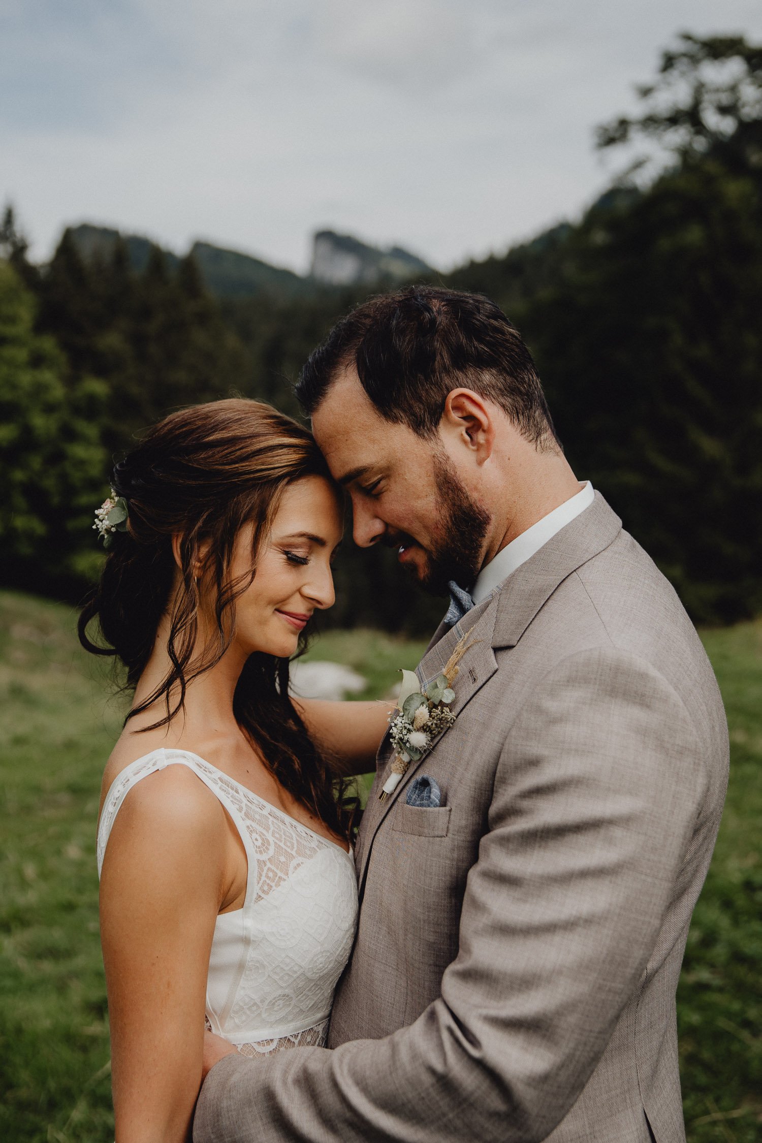 Ein Brautpaar steht im Freien, ihre Stirn und Köpfe berühren sich, beide lächeln sanft. Die Frau trägt ein weißes Hochzeitskleid, der Mann einen beigefarbenen Anzug mit Krawatte. Im Hintergrund sind Bäume und Berge sichtbar.