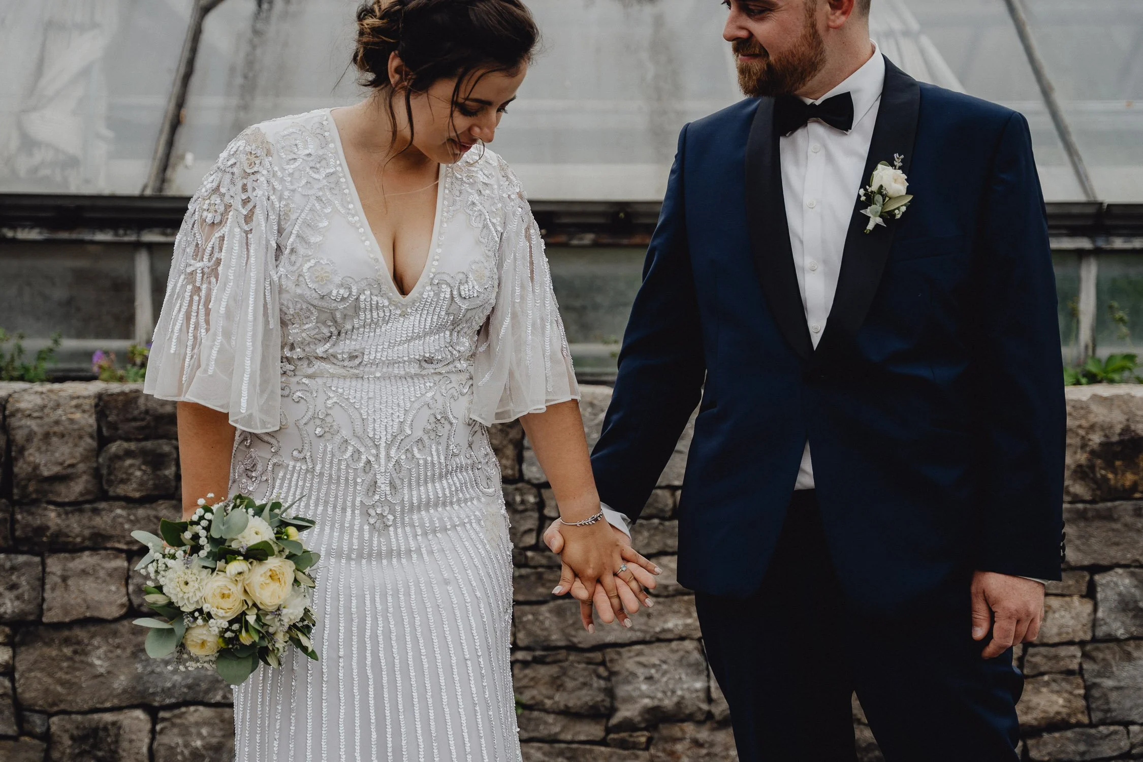 Ein Brautpaar hält Händchen bei ihrer Hochzeit; die Braut trägt ein weißes, verzierter Kleid und hält einen Blumenstrauß, der Bräutigam trägt einen schwarzen Anzug mit Fliege.