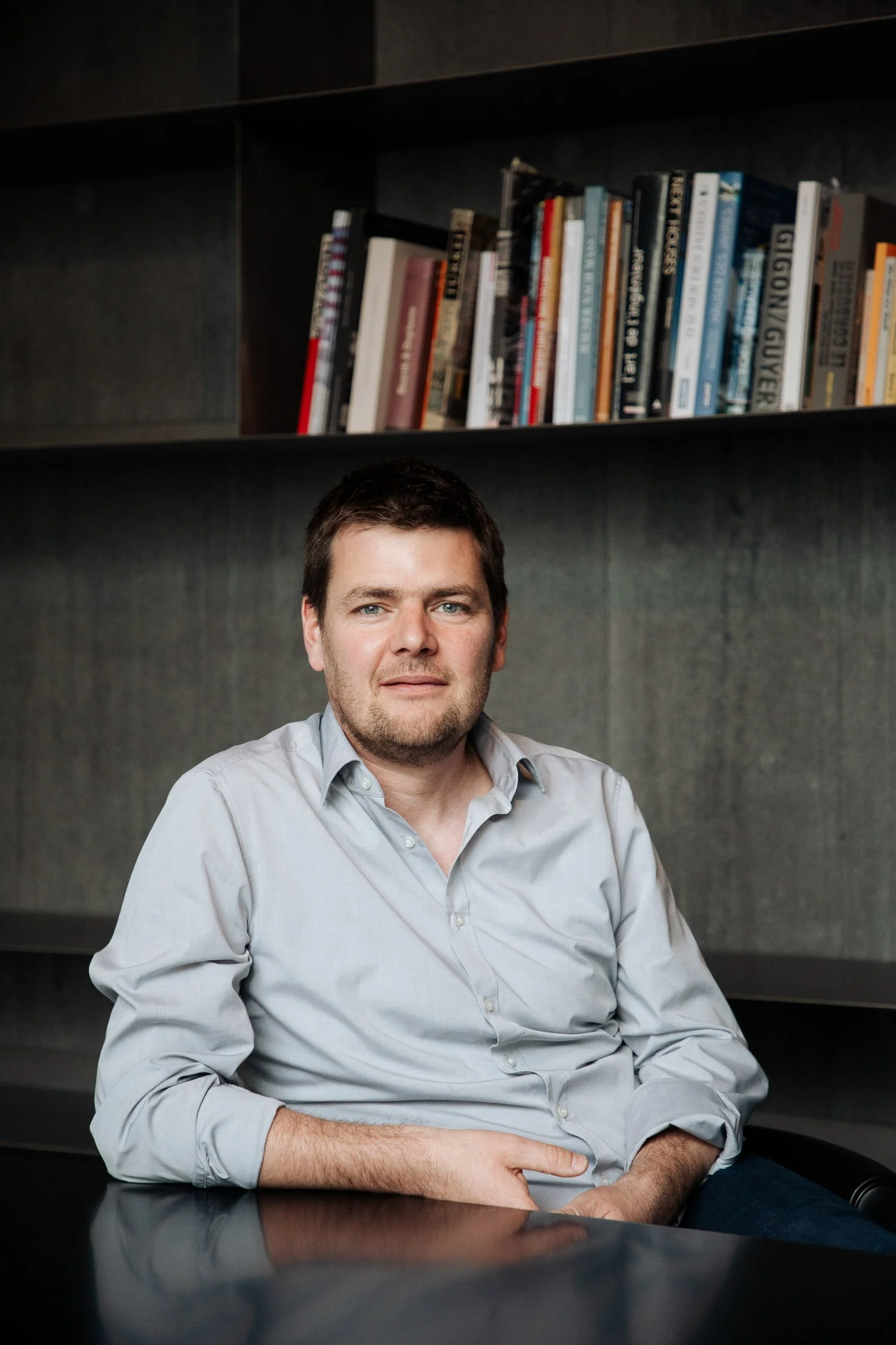 Ein Mann mit dunklem Haar und Bart sitzt an einem schwarzen Tisch vor einer Bücherregalwand.