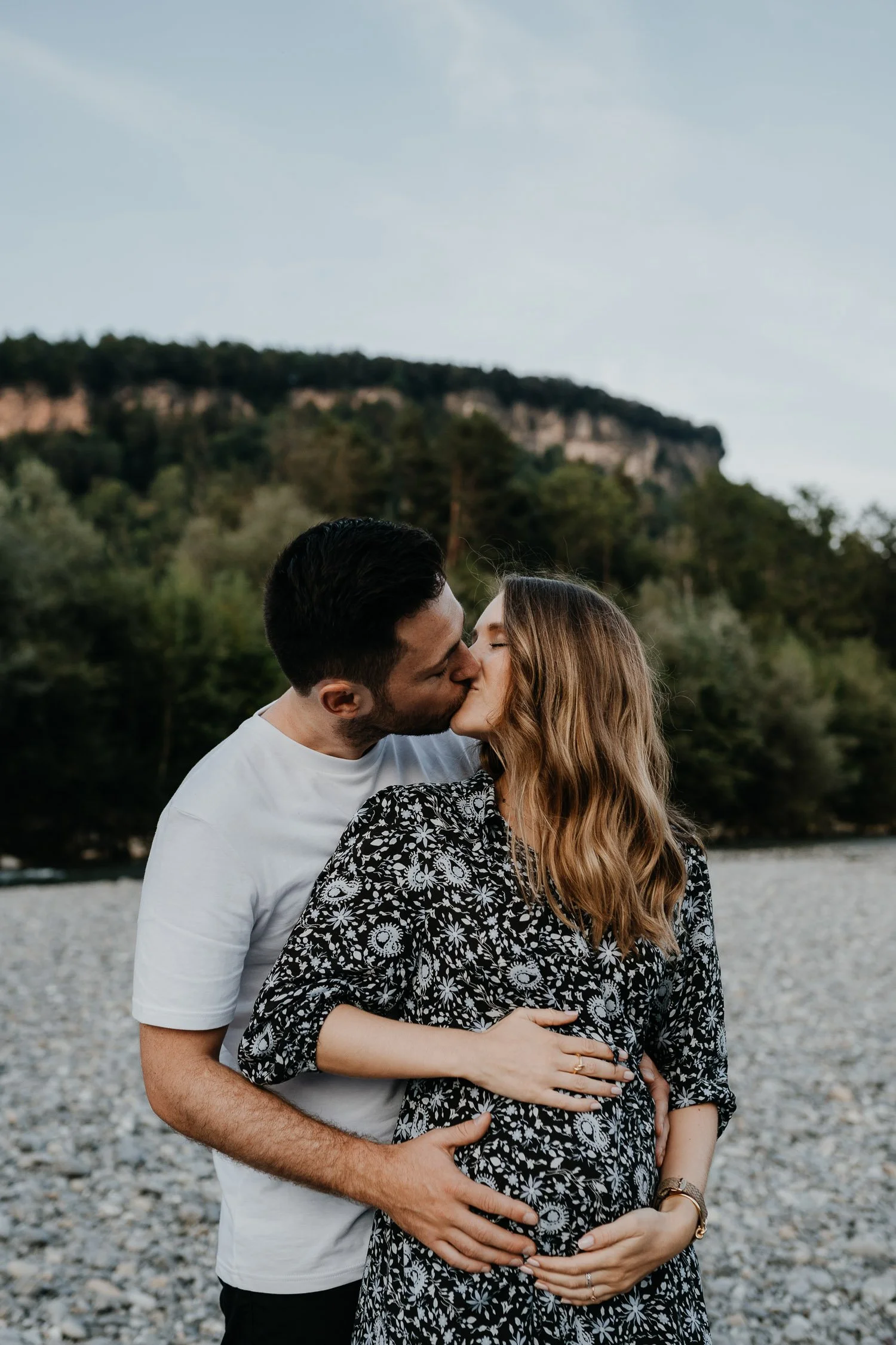 Ein schwangeres Paar küsst sich am Flussufer vor einem grünen Hügel.