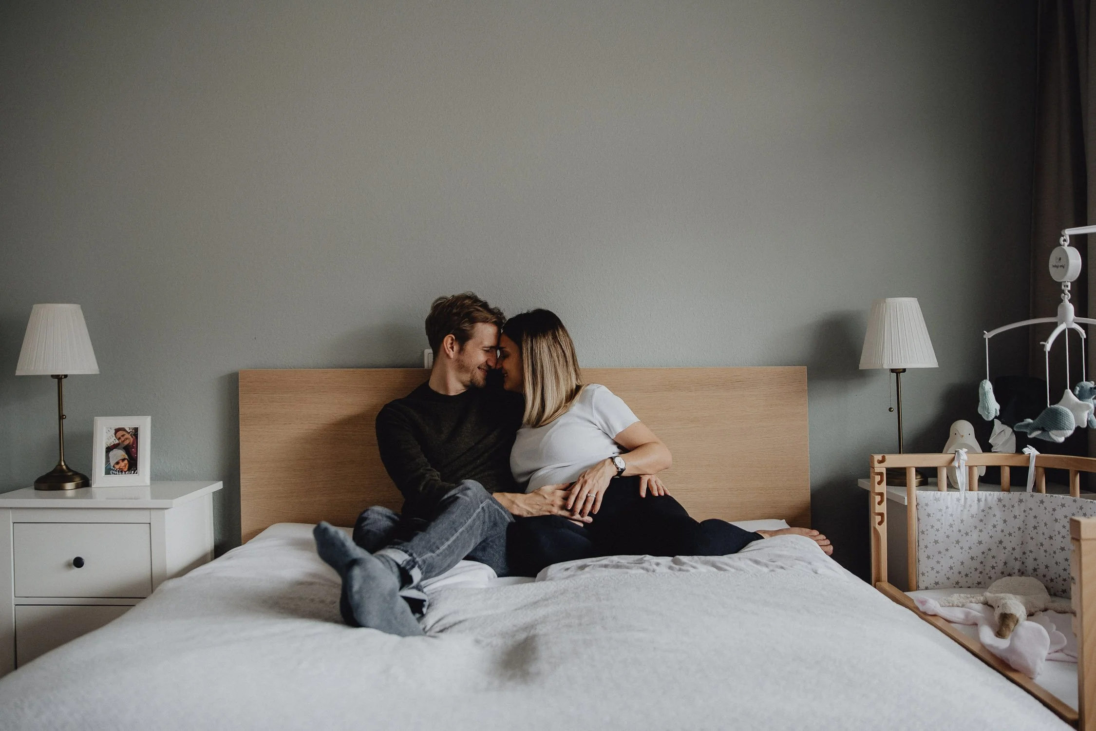 Ein Paar sitzt auf einem Bett, lächelt sich an und berührt sich liebevoll, im Schlafzimmer mit Nachttischen, Lampen und Kinderbett.