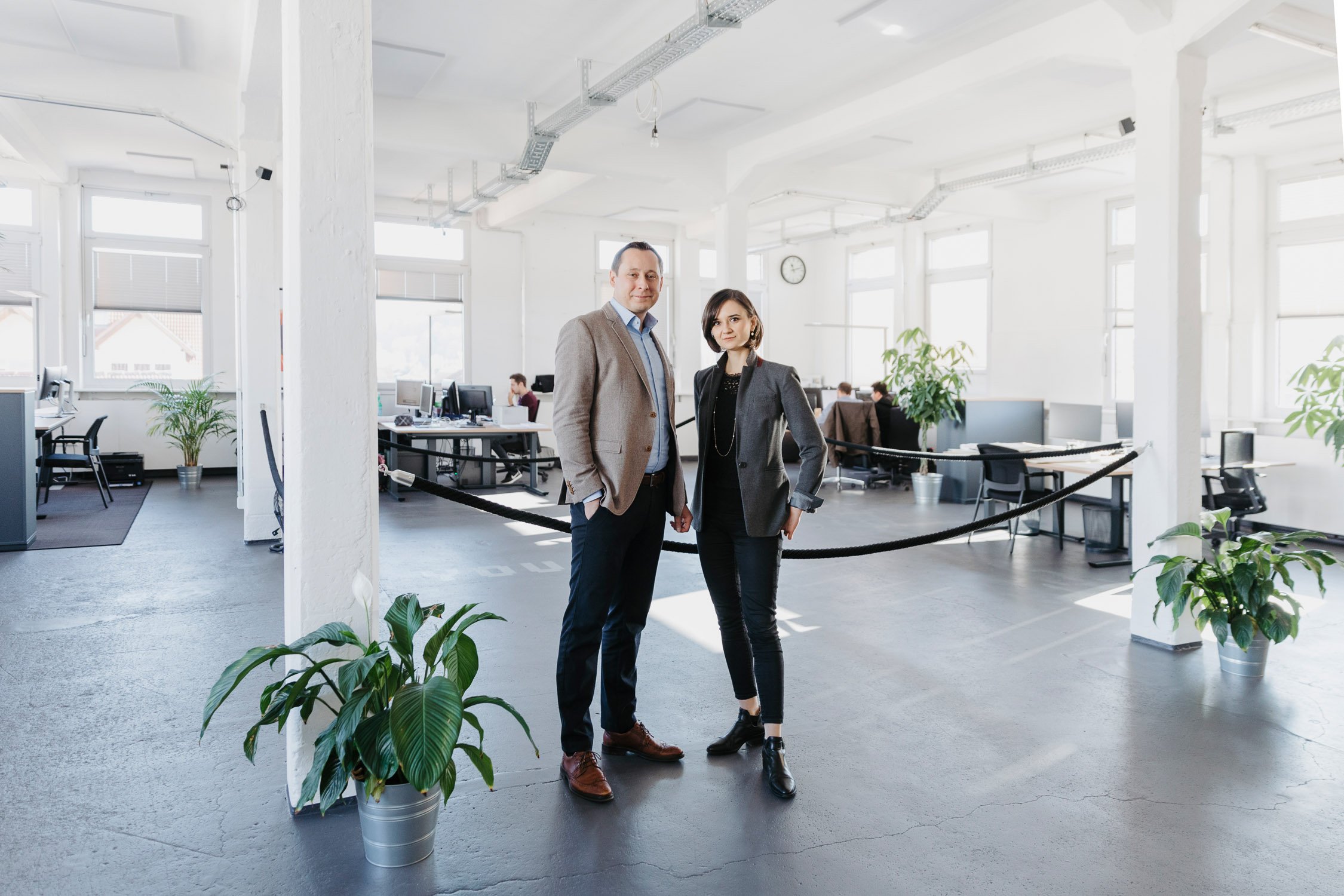Zwei berufstätige Personen, ein Mann und eine Frau, posieren im modernen Büro. Im Hintergrund sind mehrere Arbeitsplätze mit Computern und Kollegen sichtbar.