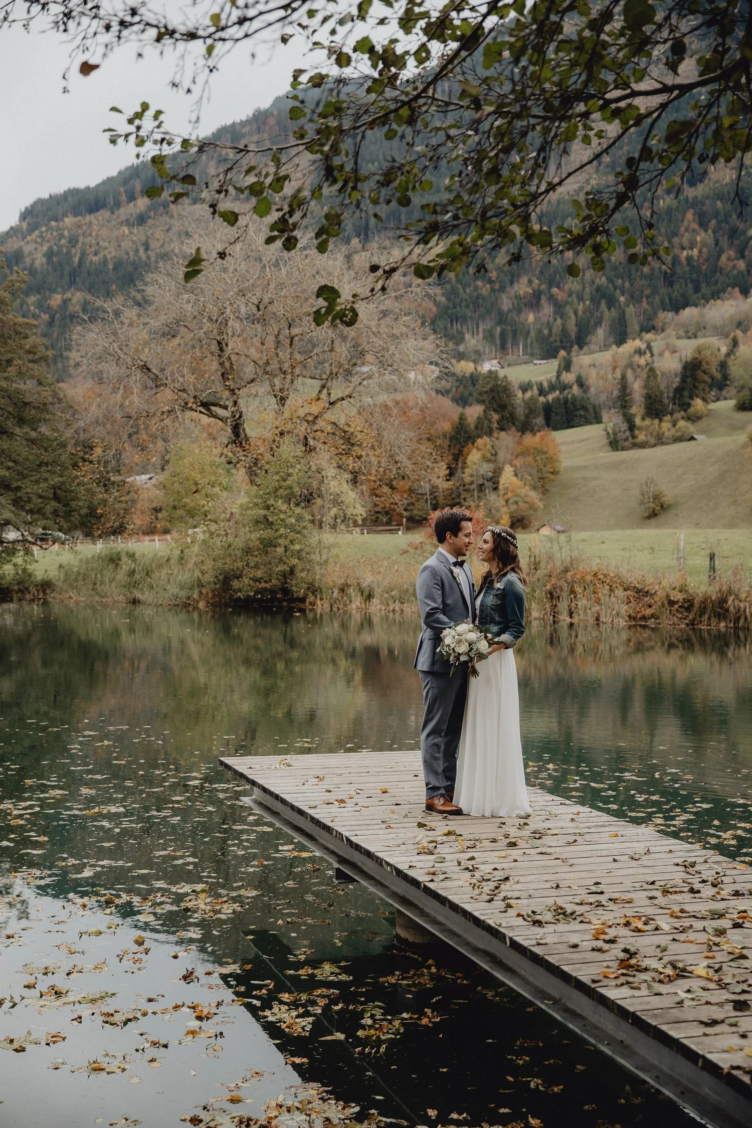 Ein Brautpaar steht auf einem Steg an einem See in einer malerischen Herbstlandschaft, umgeben von Bäumen mit goldenem und orangefarbenem Laub, im Hintergrund Berge. Das Paar schaut sich verliebt an, die Braut trägt ein langes weißen Kleid und hält e