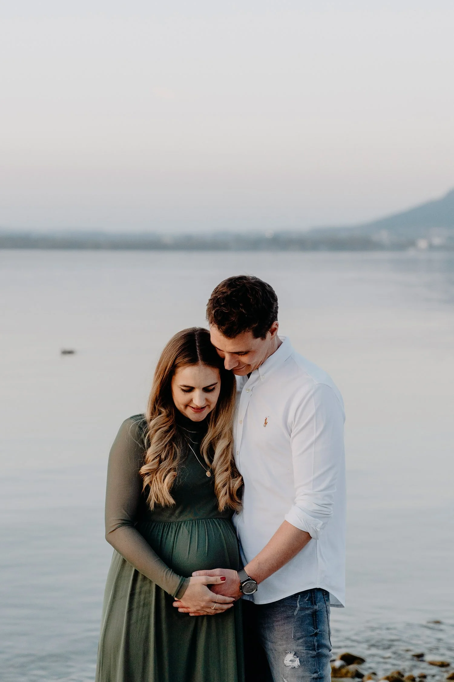 Ein Paar steht am Flussufer, der schwangere Frau trägt ein langes grünes Kleid, der Mann trägt ein weißes Hemd, beide halten die Hände an ihrem Babybauch, bei Sonnenuntergang
