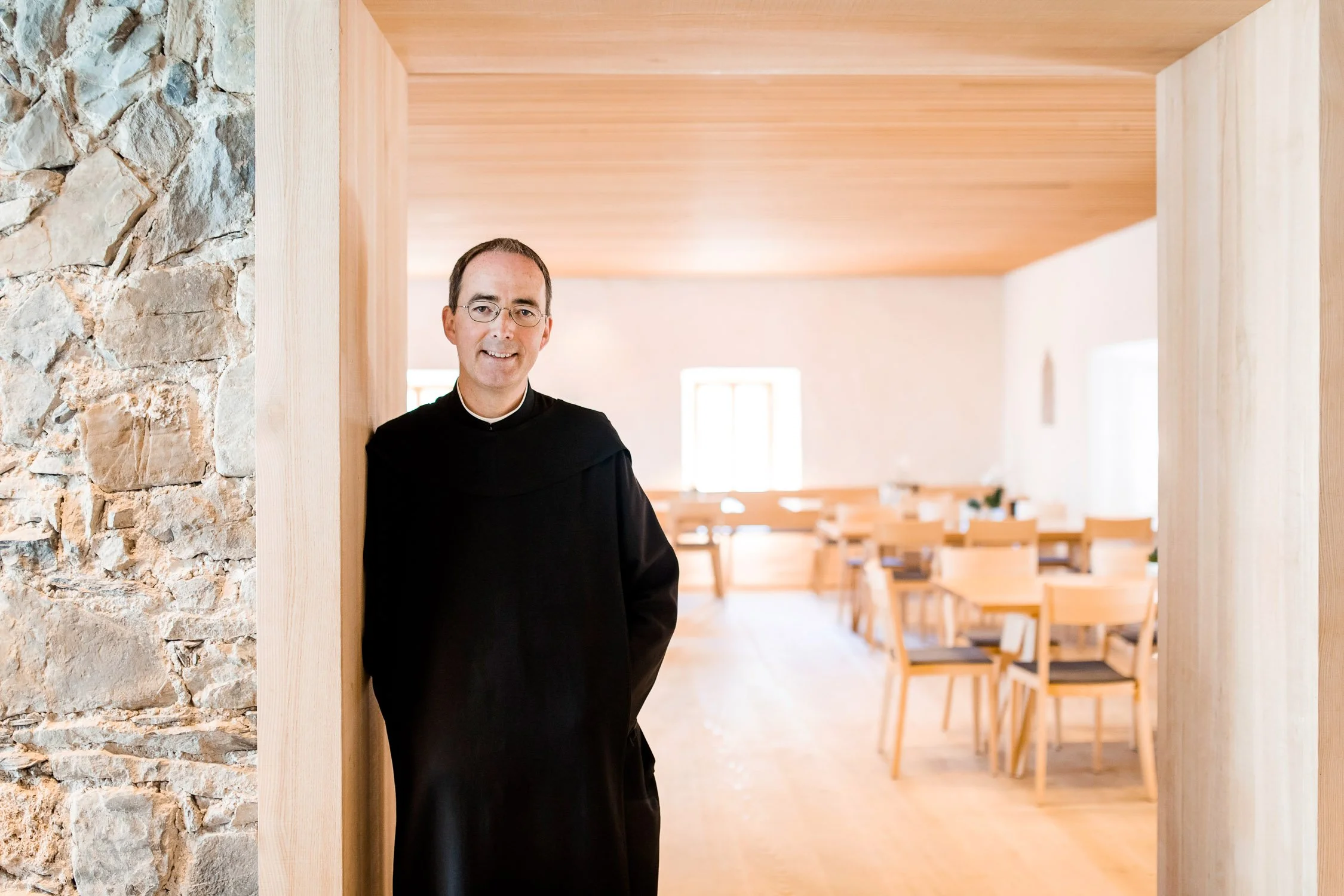 Ein Priester steht in einem Kirchenraum mit hellem Holzdekor, Blick durch einen Türrahmen, im Hintergrund leere Stühle.