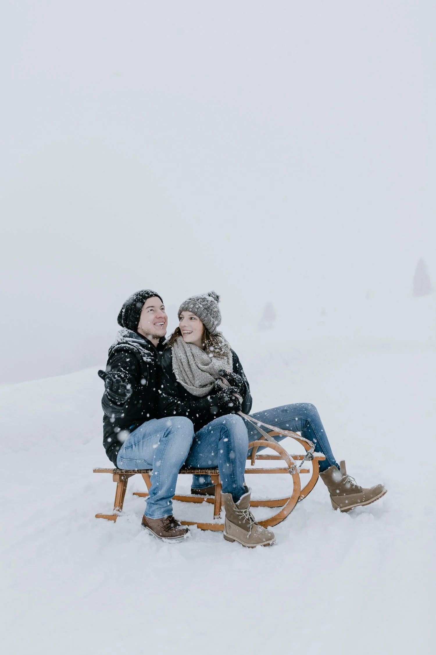 Ein Paar sitzt auf einer Schlitten im Schnee, umgeben von Schneefall, mit winterlicher Kleidung und lachend.