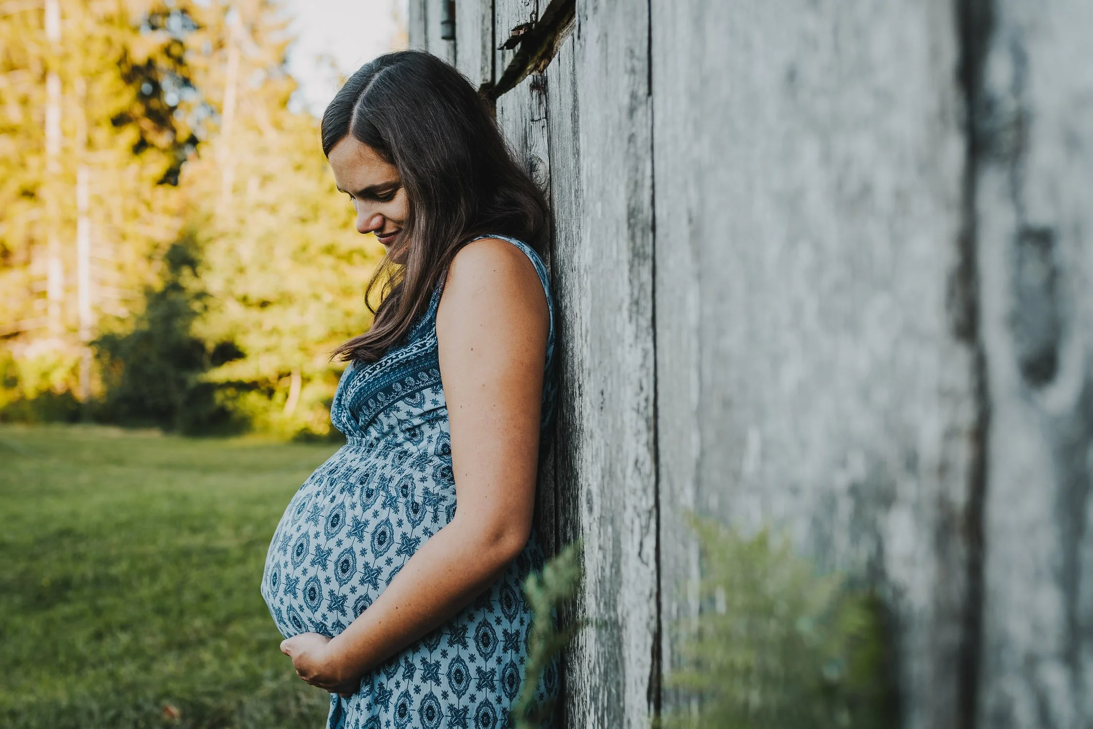 Schwangere Frau lehnt an einer Holzwand im Freien, umgeben von Bäumen und Grünflächen, lächelnd nach unten.