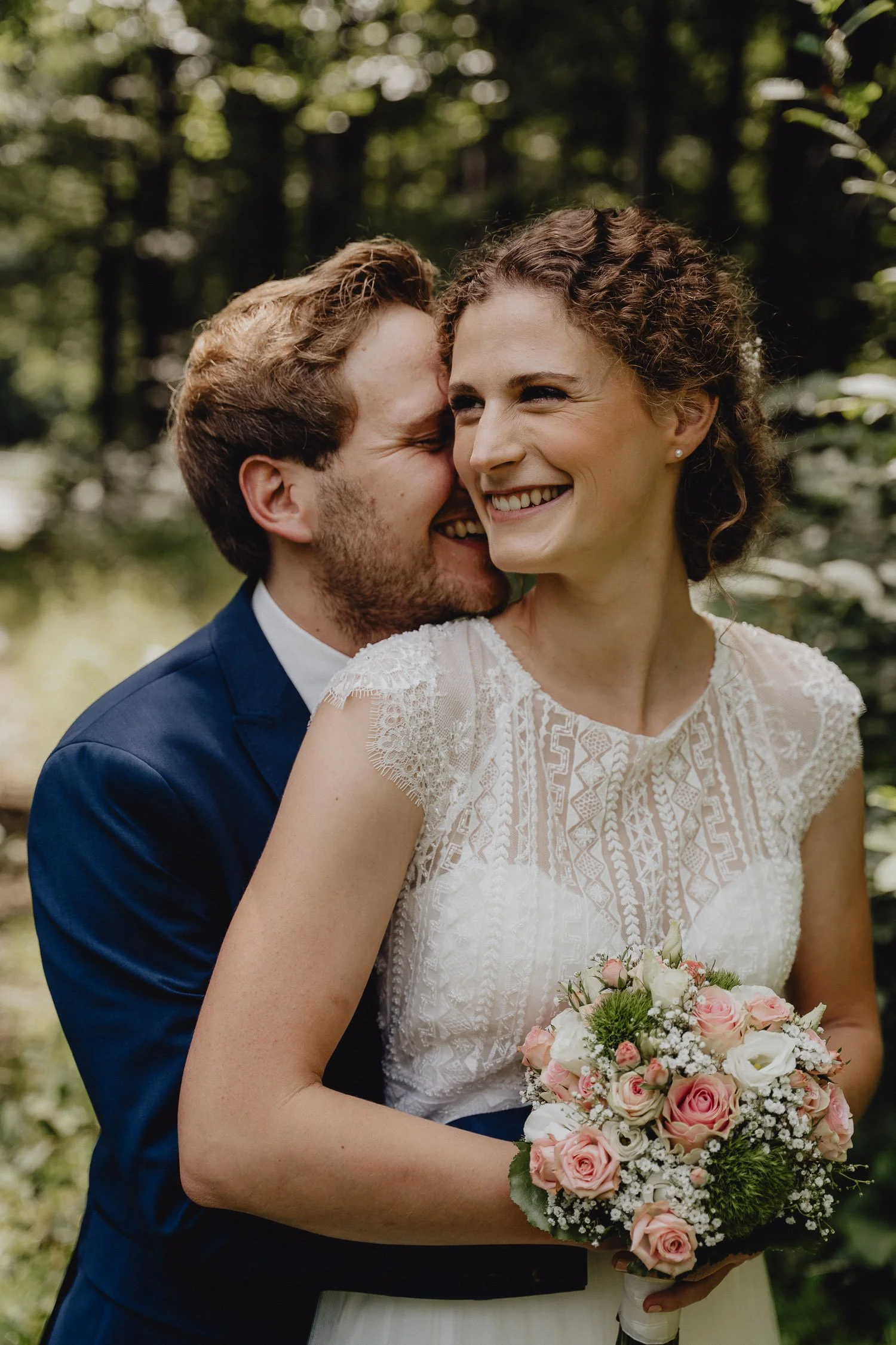 Ein lächelndes Brautpaar bei einer Hochzeit im Freien, die Frau hält einen Blumenstrauß mit rosa und weißen Rosen, im Hintergrund sind grüne Bäume.