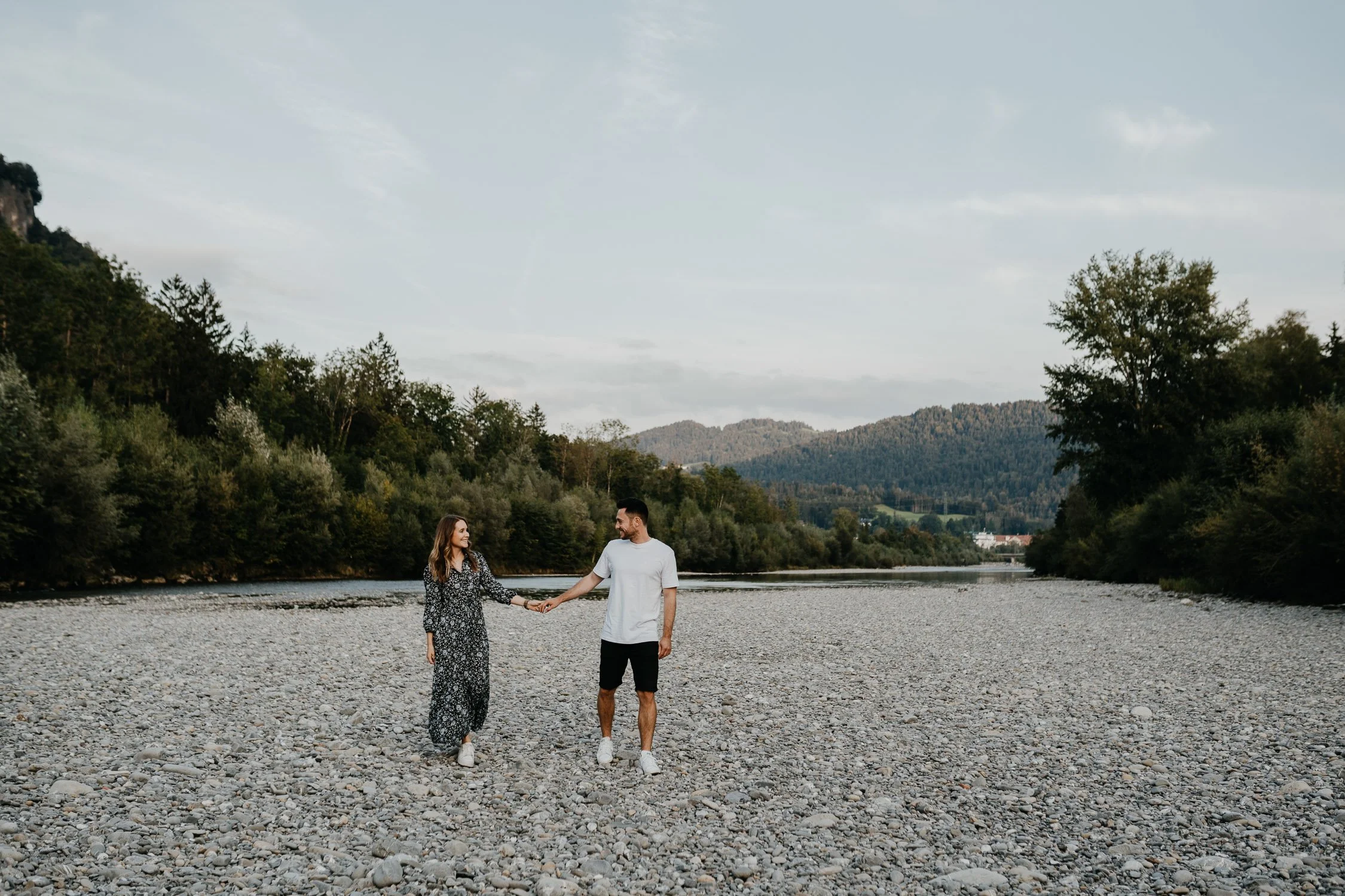 Ein Paar hält sich an einem Flussufer mit Kiessteinboden, umgeben von grünen Bäumen und Bergen im Hintergrund, bei Sonnenuntergang.