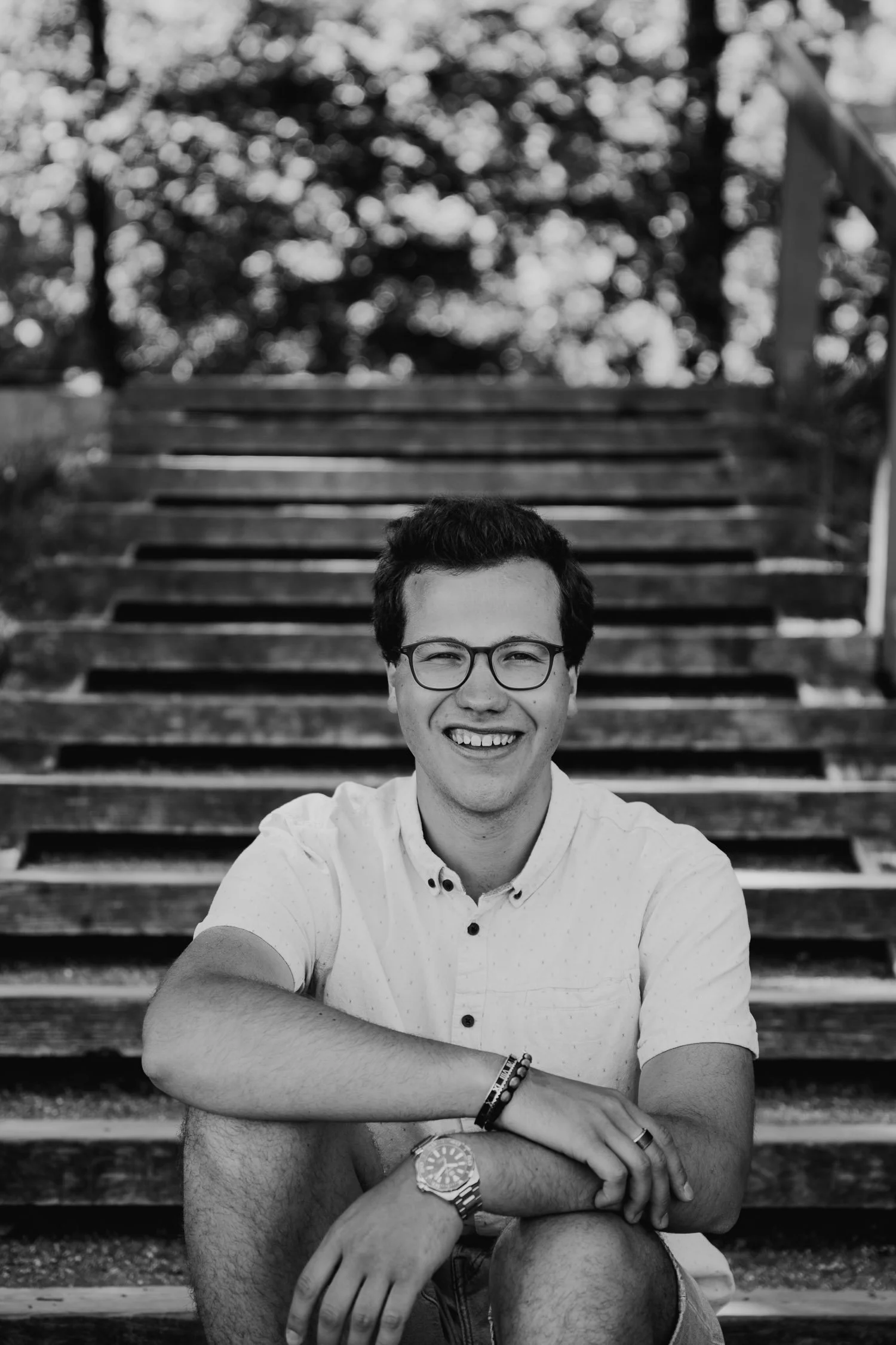 Junge Mann sitzt auf den Holzstufen im Freien, lächelt in die Kamera, trägt Brille, weißes Hemd und Armband, schwarze Uhr, Bokeh-Hintergrund mit Bäumen.