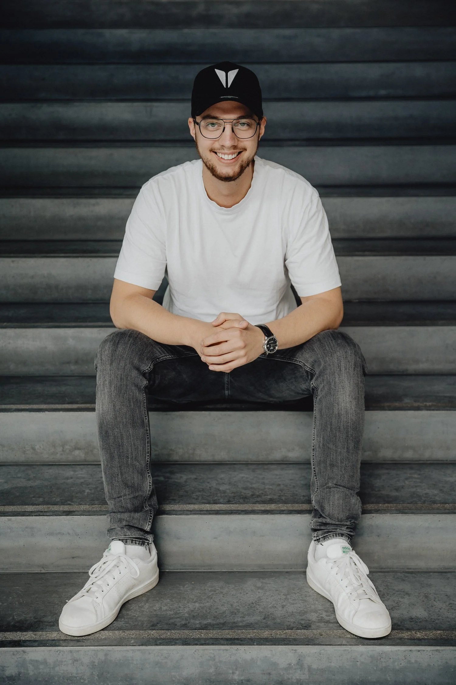 Junger Mann sitzt auf Treppen, trägt eine weiße T-Shirt, graue Jeans, weiße Sneakers, eine schwarze Baseballkappe und eine Armbanduhr, lächelt