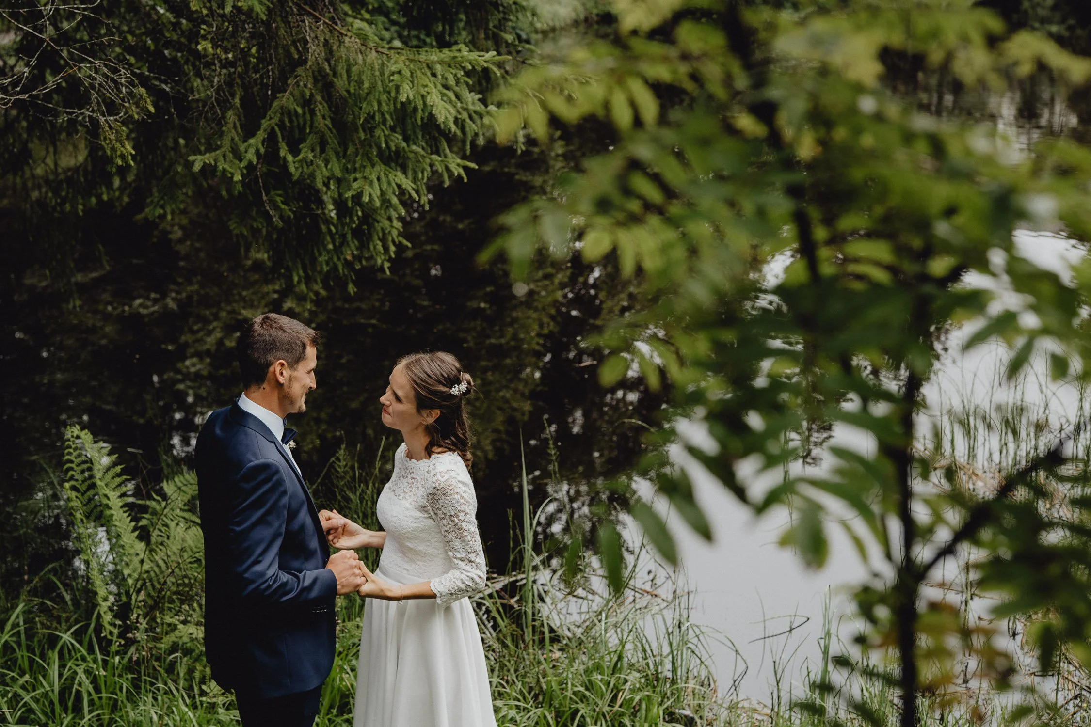 Ein Brautpaar steht an einem See im Grünen, hält Händchen und schaut sich liebevoll an, umgeben von Bäumen und Wasser.